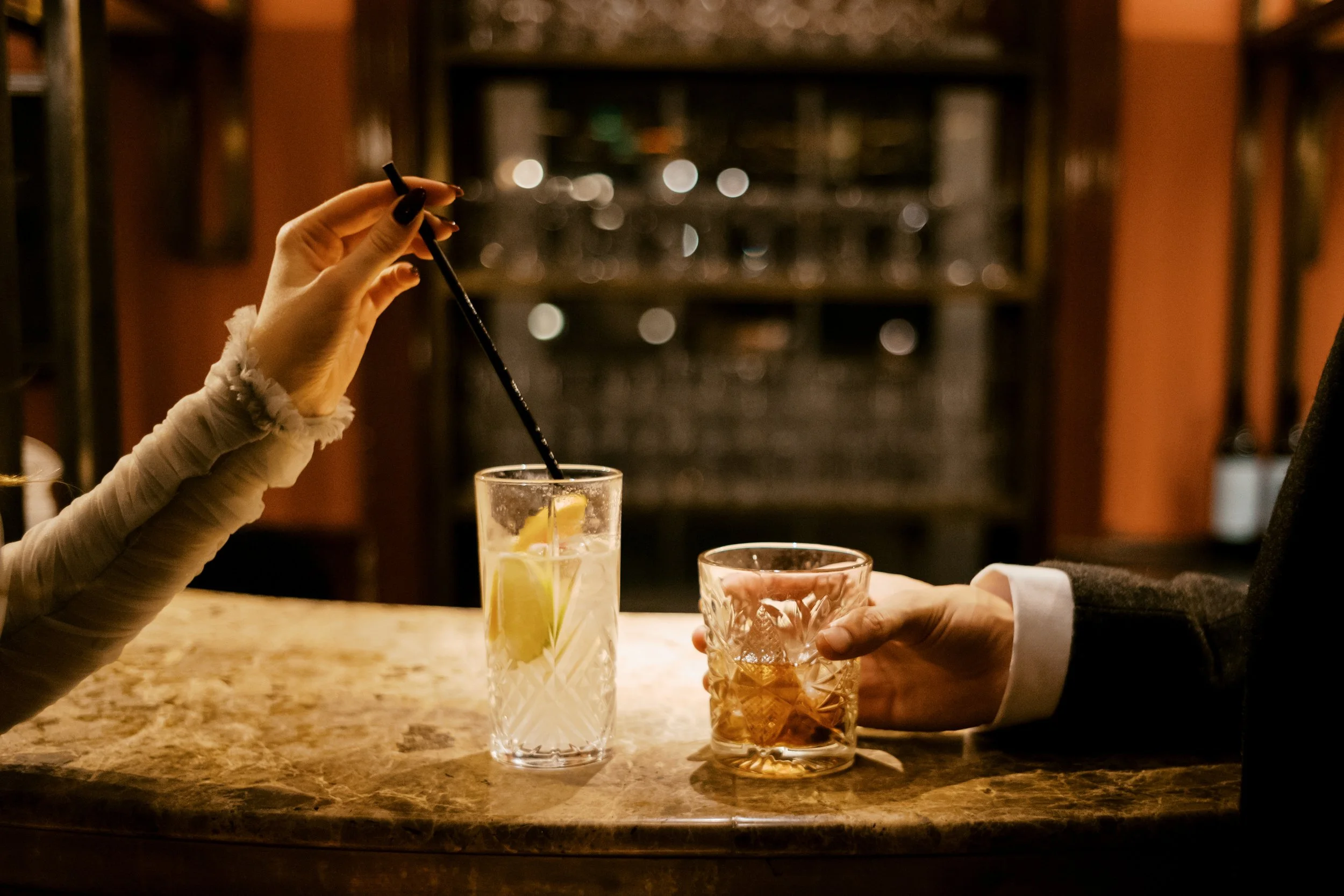 Person with a black painted fingernail stirring a lemon slice drink with a black straw at a bar, while another person holds a glass containing ice and a brown drink, on a marble bar counter.