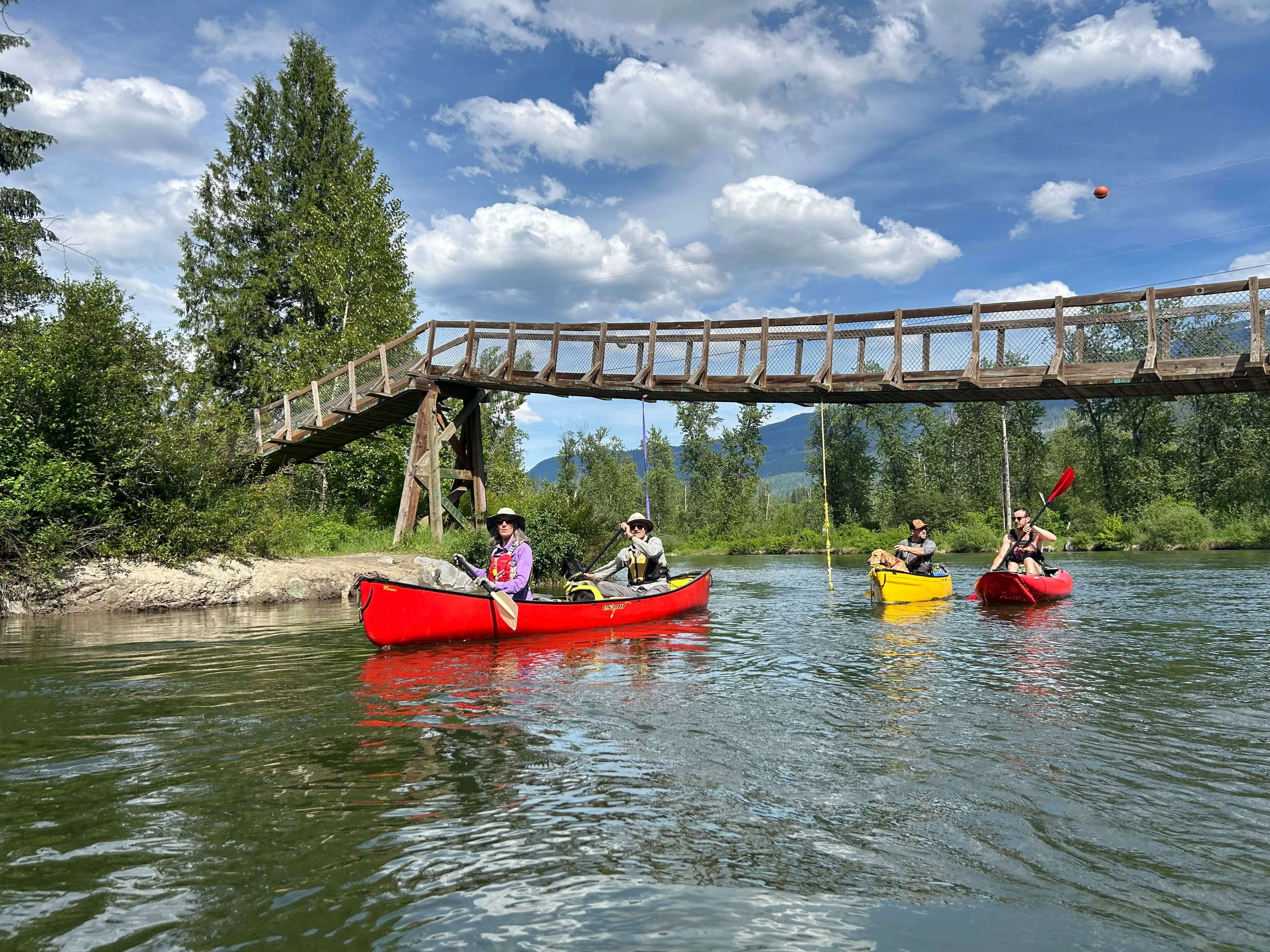 People canoeing on a river beneath a wooden footbridge with a chain-link fence, surrounded by lush green trees and a partly cloudy sky.
