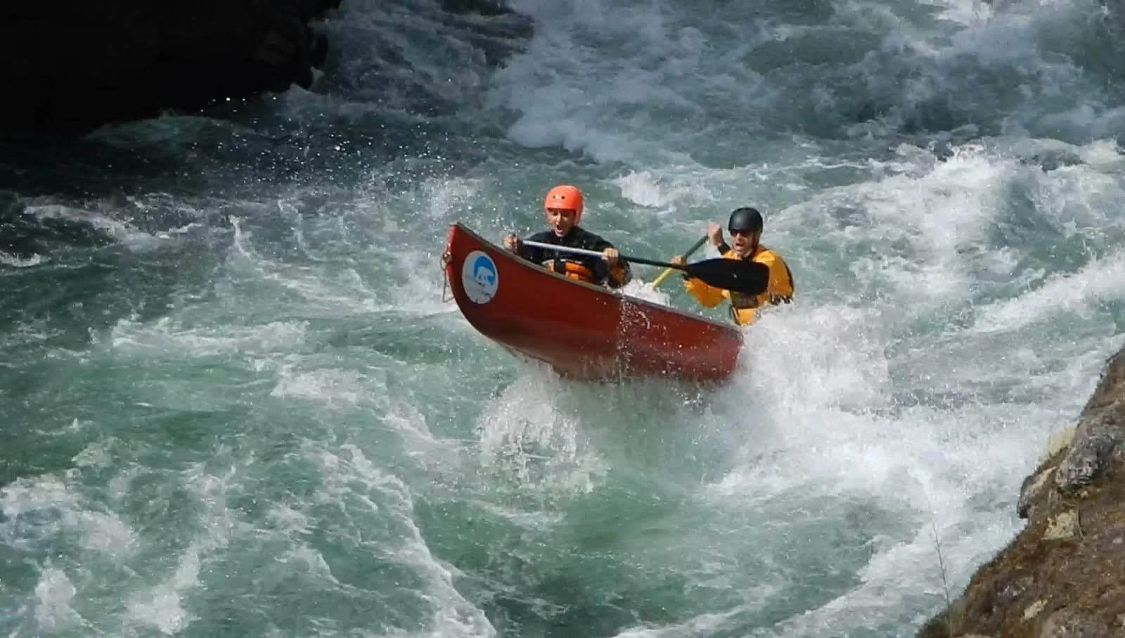 Two people in a red kayak navigating turbulent white water rapids, wearing helmets and life jackets, with rocky banks on either side.