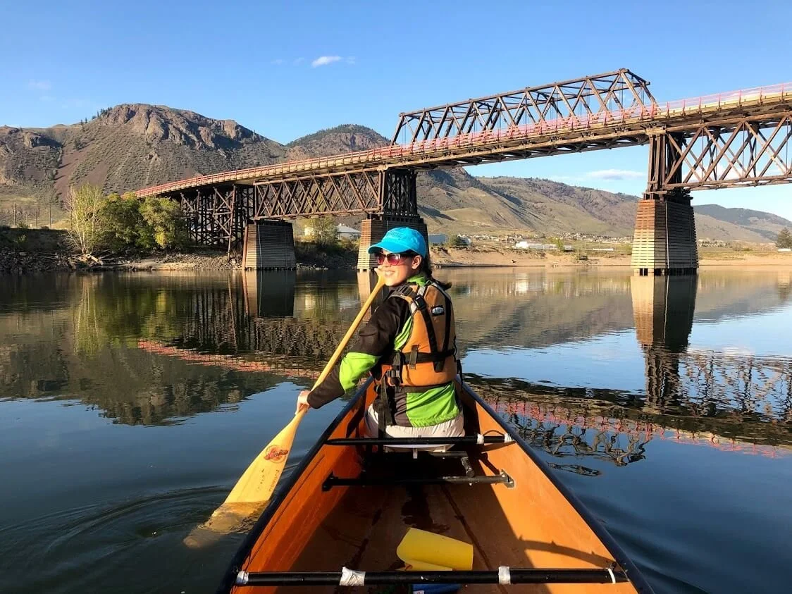 A woman paddling on a calm river with mountains and a bridge in the background