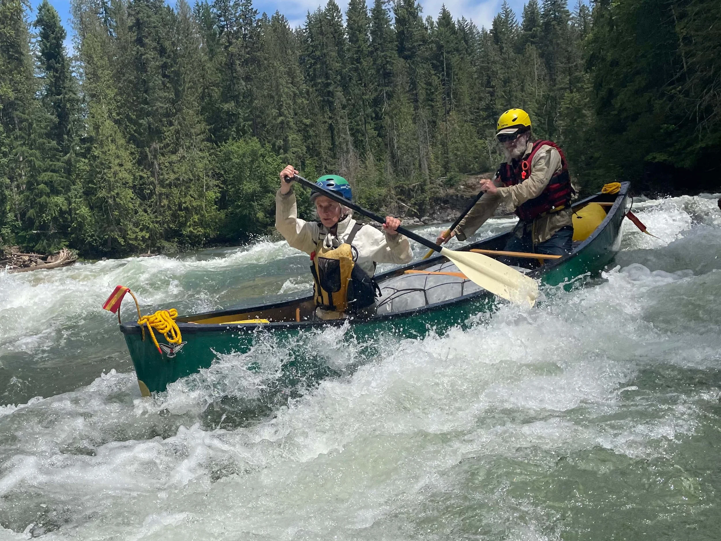 Two people white water canoeing on a river surrounded by dense green forest, both wearing helmets and life jackets, with one person paddling at the front and the other paddling at the back.