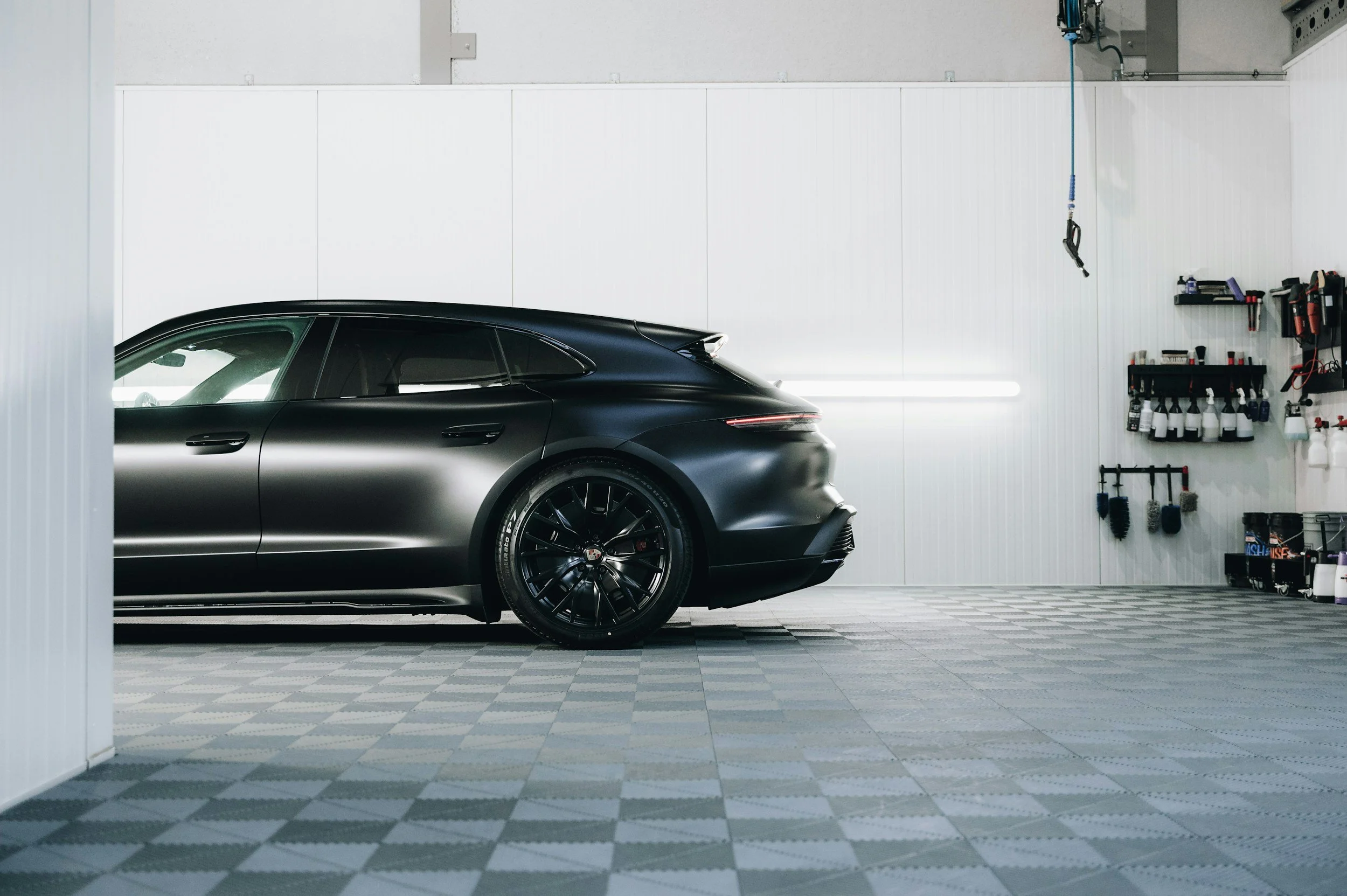 Black luxury sports car parked inside a white-walled garage with tools and supplies on shelves on the right.