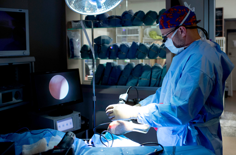 Surgeon in scrubs and a mask performing a procedure in an operating room with medical equipment and supplies.