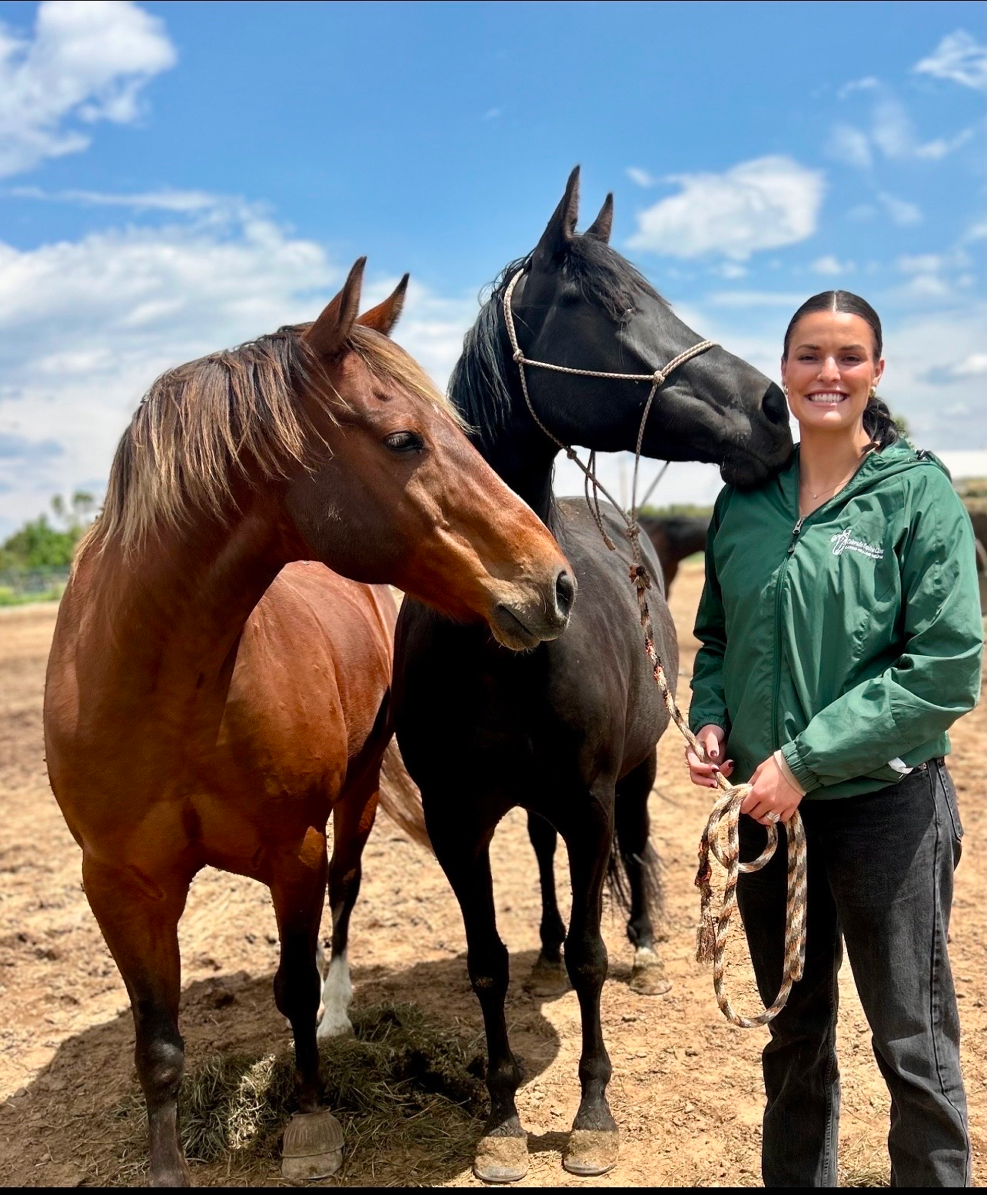 A woman standing outdoors in a green jacket, smiling, with three horses on a cloudy day.