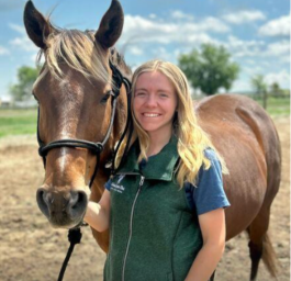 A young woman with blonde hair smiling next to a brown horse outdoors on a sunny day.