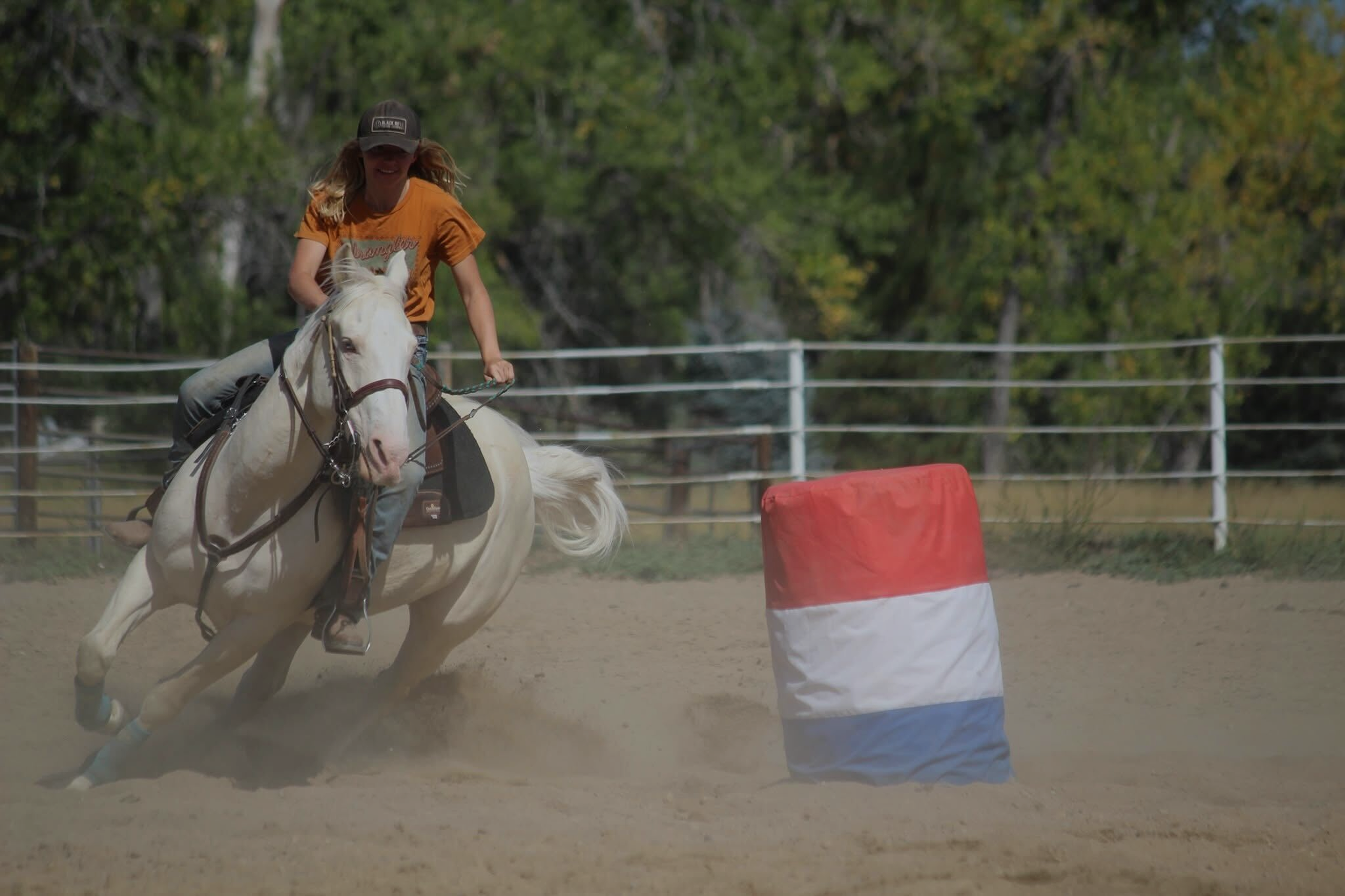 A woman riding a white horse on a dirt track during a horse riding event, with a red and white barrier in front of her and a white fence behind, surrounded by trees.