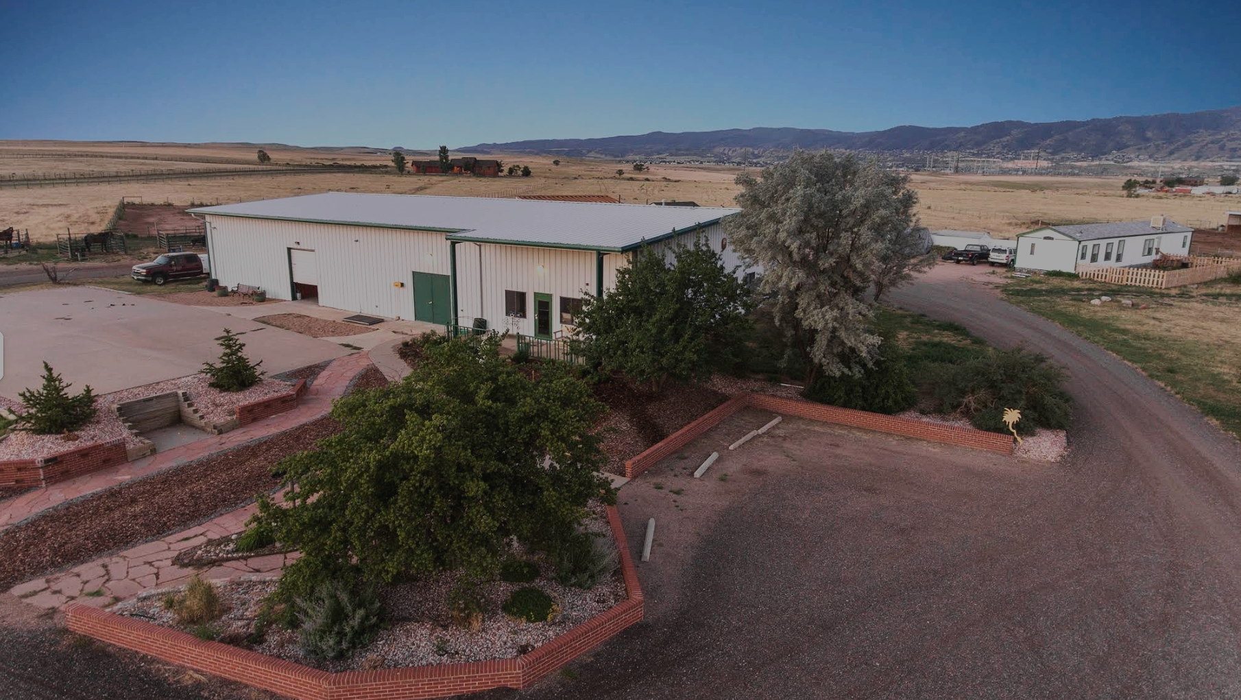 Aerial view of a rural farm with a large white barn, surrounding trees, gravel driveway, parked trucks, and distant open fields with mountains in the background.