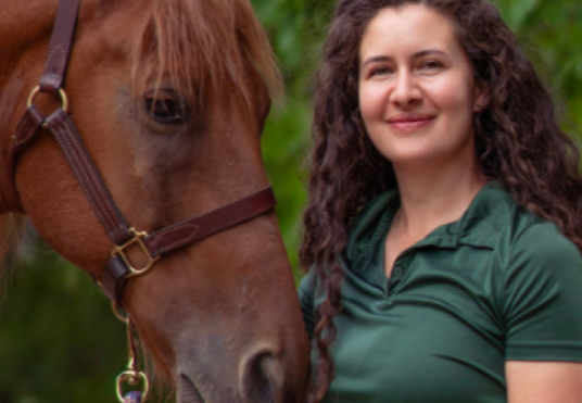 A woman with curly dark hair standing next to a brown horse with a halter, outdoors with green foliage in the background.
