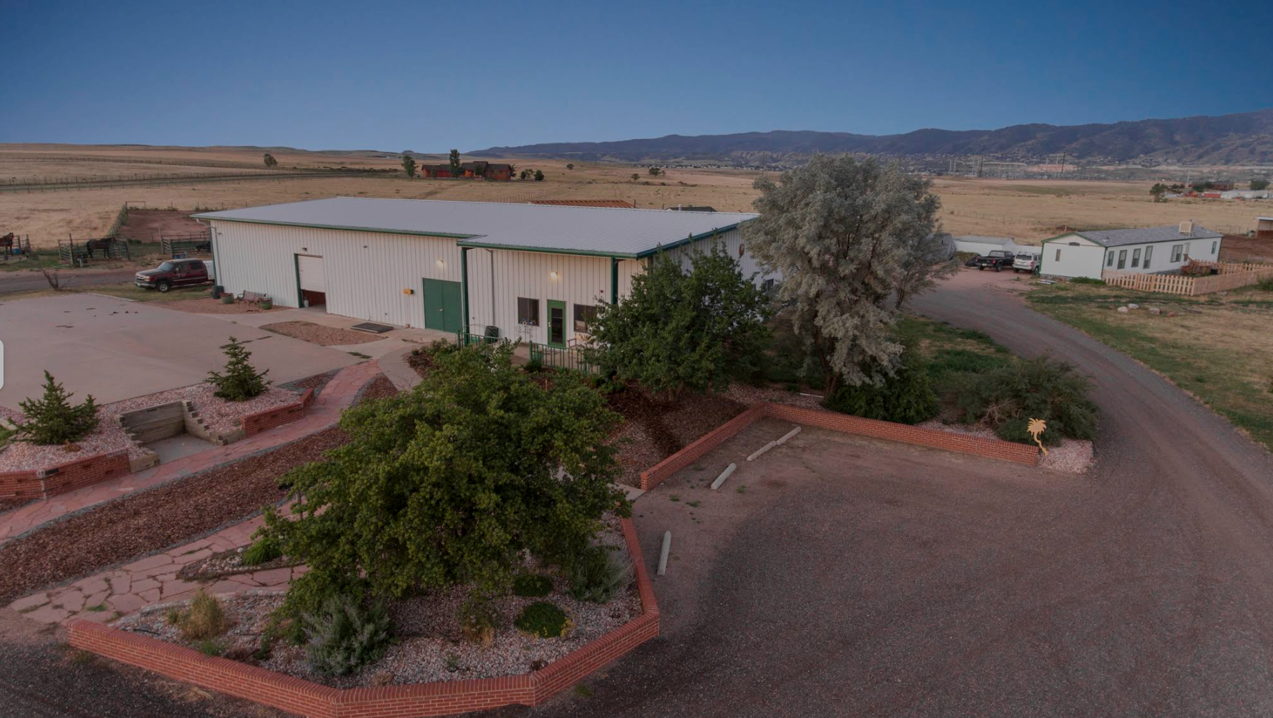 A rural scene at dusk featuring a large white metal building with a green door, surrounded by landscaped yards with brick borders, trees, and parking areas with several vehicles. The background includes open fields, distant hills, and a clear evening sky.