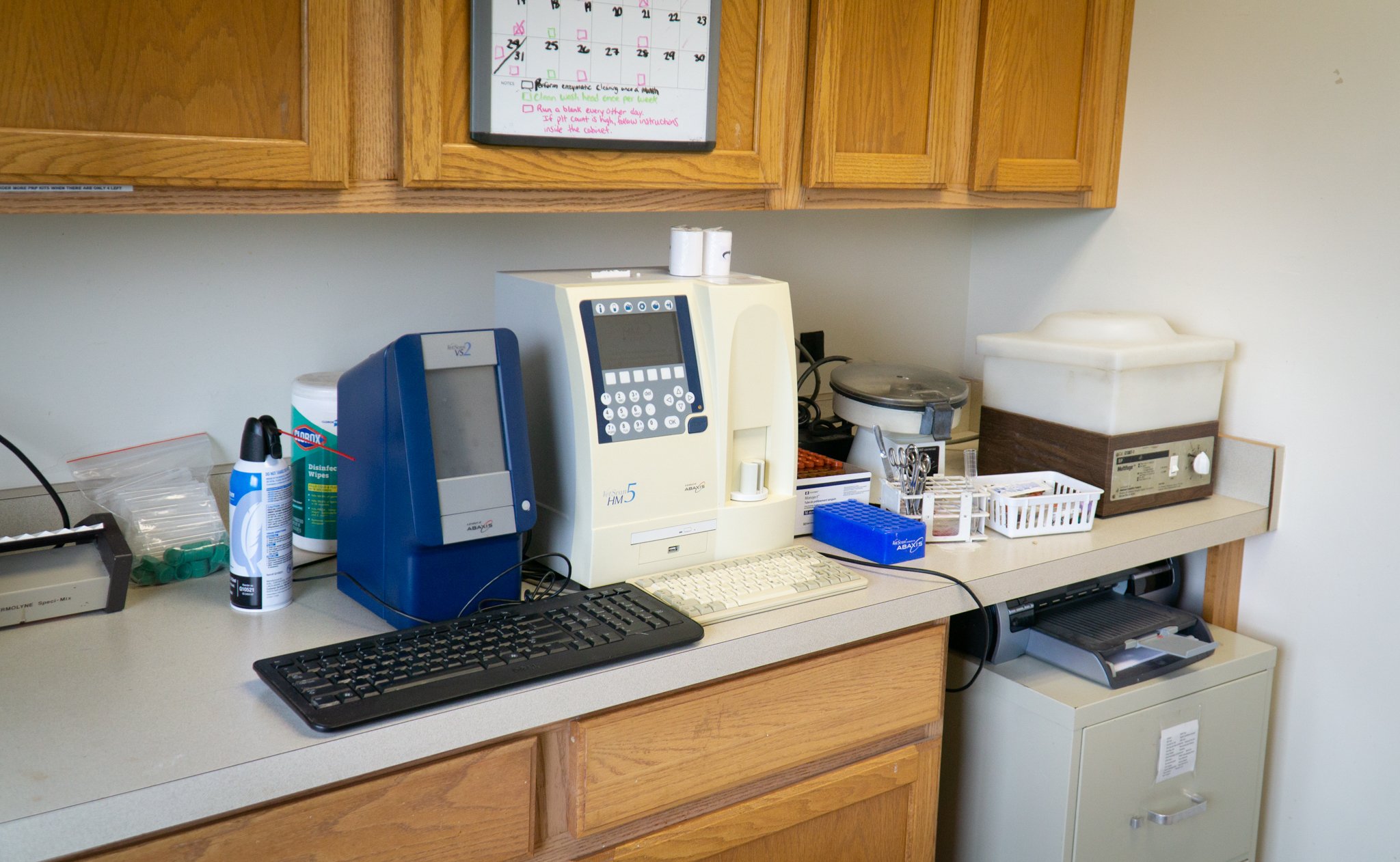 Medical or laboratory workspace with equipment including a computer, printer, and various supplies organized on a countertop with wooden cabinets above.