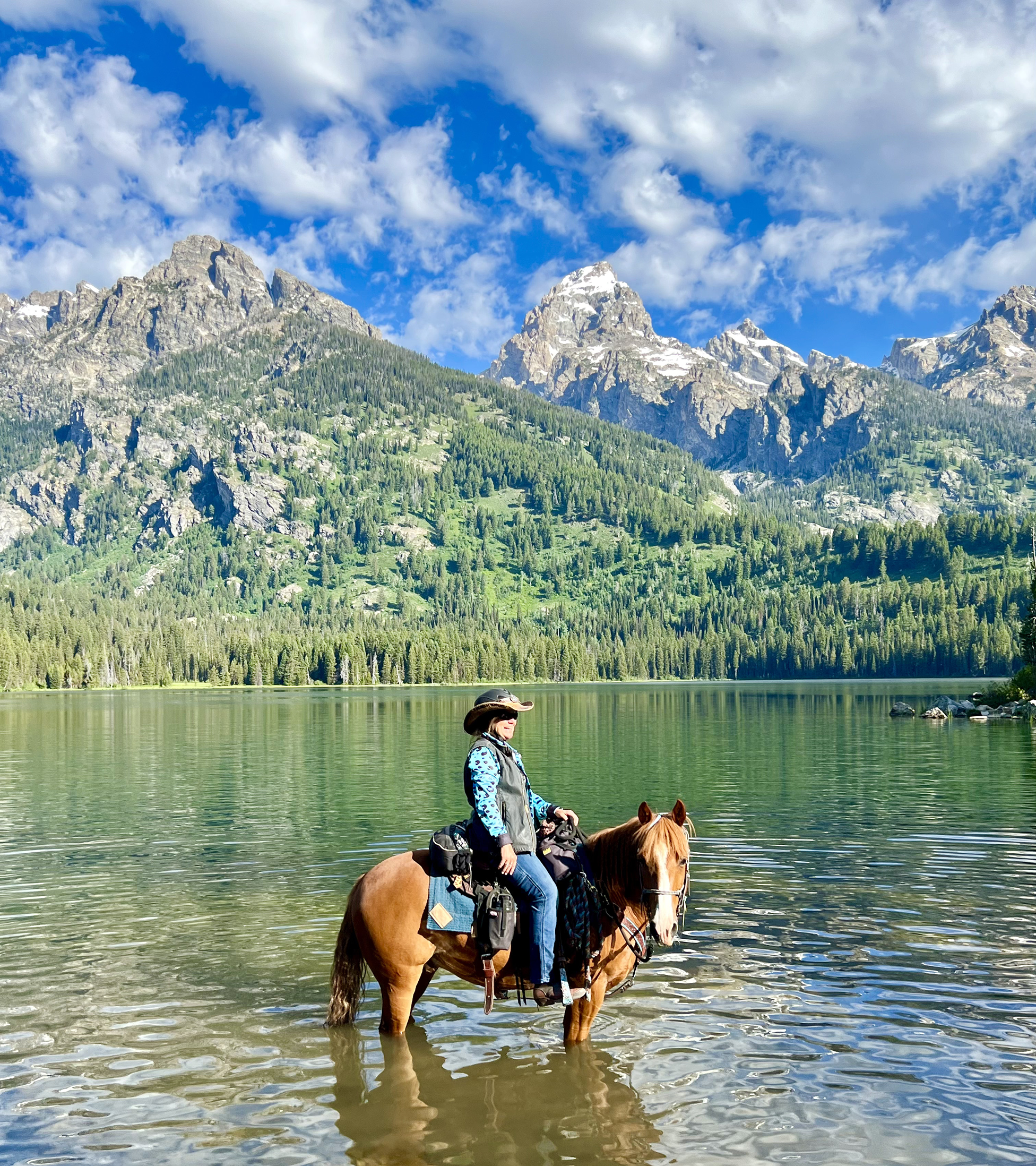 A woman on horseback in a lake with a mountain range and a blue sky with clouds in the background.