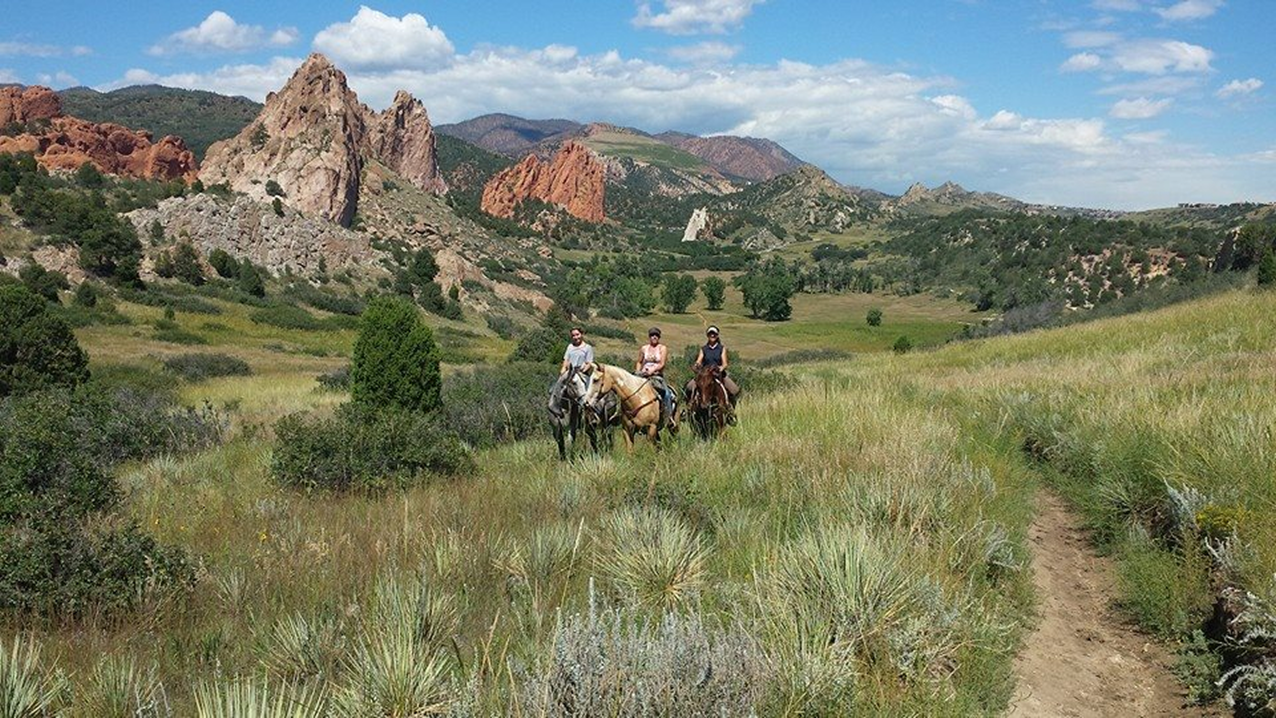 Three people riding horses through a grassy field with mountains and a partly cloudy sky in the background.