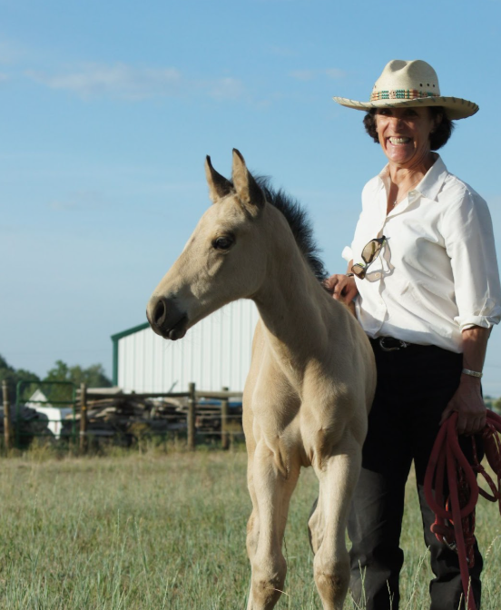 Woman smiling and standing next to a light-colored foal on a grassy field with a barn in the background.