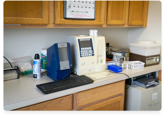 Medical laboratory equipment on a countertop with cabinets above, including an automated blood analyzer, a barcode scanner, and various supplies.