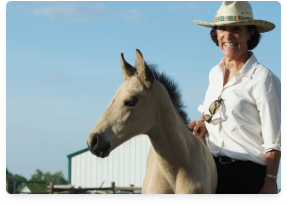 A woman smiling and wearing a wide-brimmed hat, sunglasses hanging on her shirt, sitting on a light-colored horse outdoors with a blue sky and a barn in the background.