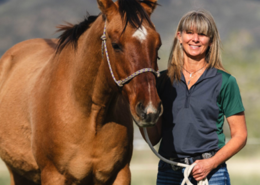 A woman with blonde hair and a blue and green shirt standing next to a brown horse with a white stripe on its face in an outdoor setting.