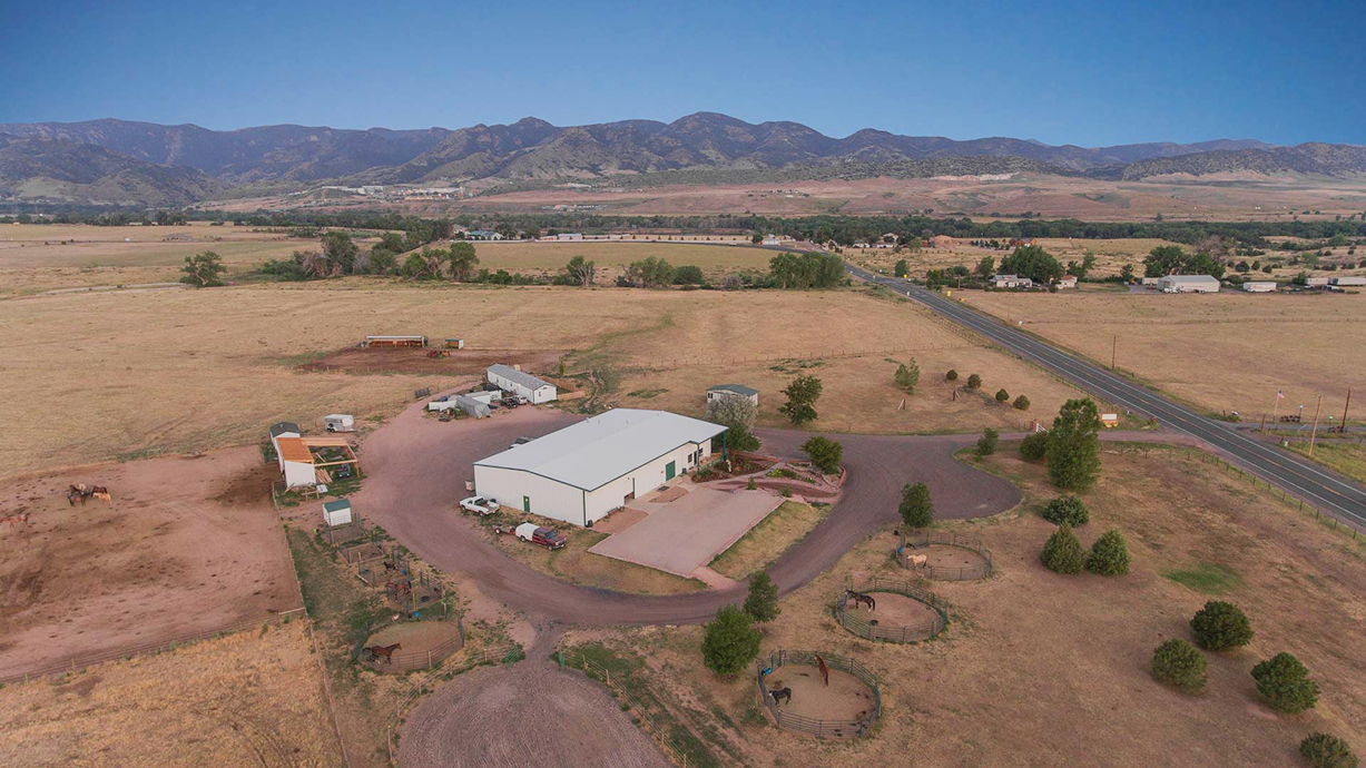Aerial view of a farm with a large white building, horse paddocks, trees, and open fields with mountains in the background.
