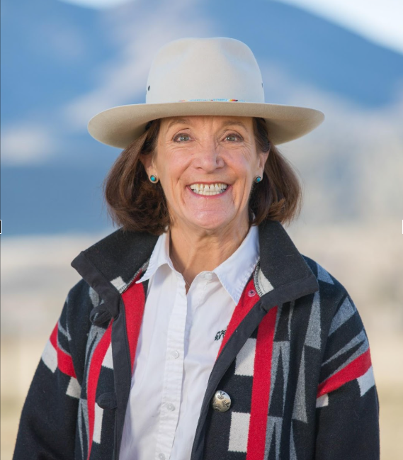 Smiling woman wearing a white hat, white shirt, and patterned jacket outdoors with mountains in the background.