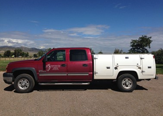 Red pickup truck with utility bed parked on gravel under a partly cloudy blue sky with mountains and trees in the background.