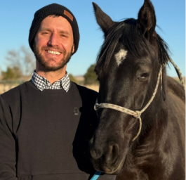 A smiling man wearing a black beanie standing next to a black horse with a white star on its forehead against a clear blue sky.