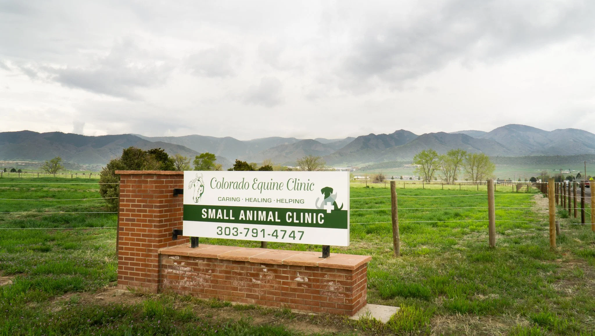 Sign for Colorado Equine Clinic small animal clinic with phone number, set against a rural landscape with mountains, fields, and trees under a cloudy sky.