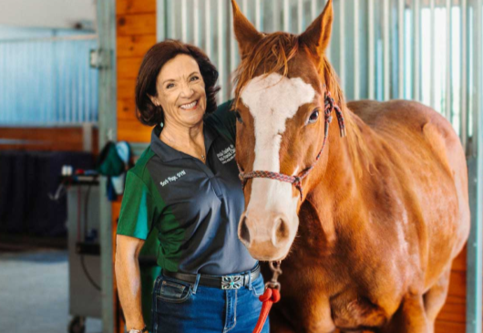 A woman standing next to a horse inside a stable, smiling at the camera.
