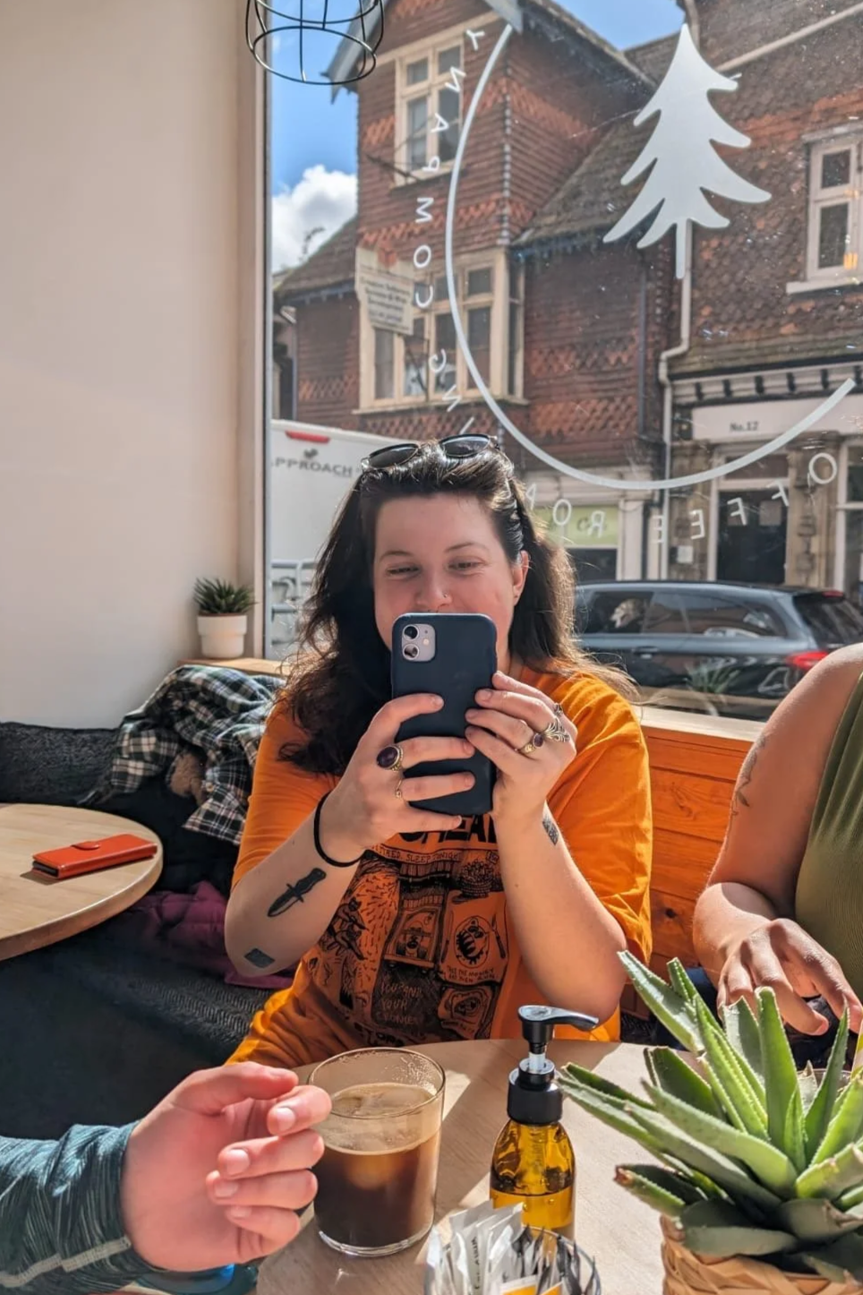 A woman with dark curly hair taking a selfie with her phone inside a cafe, sitting at a wooden table with a glass of dark coffee, a succulent plant, and a bottle of hand sanitizer, with a sunny street view outside the window.