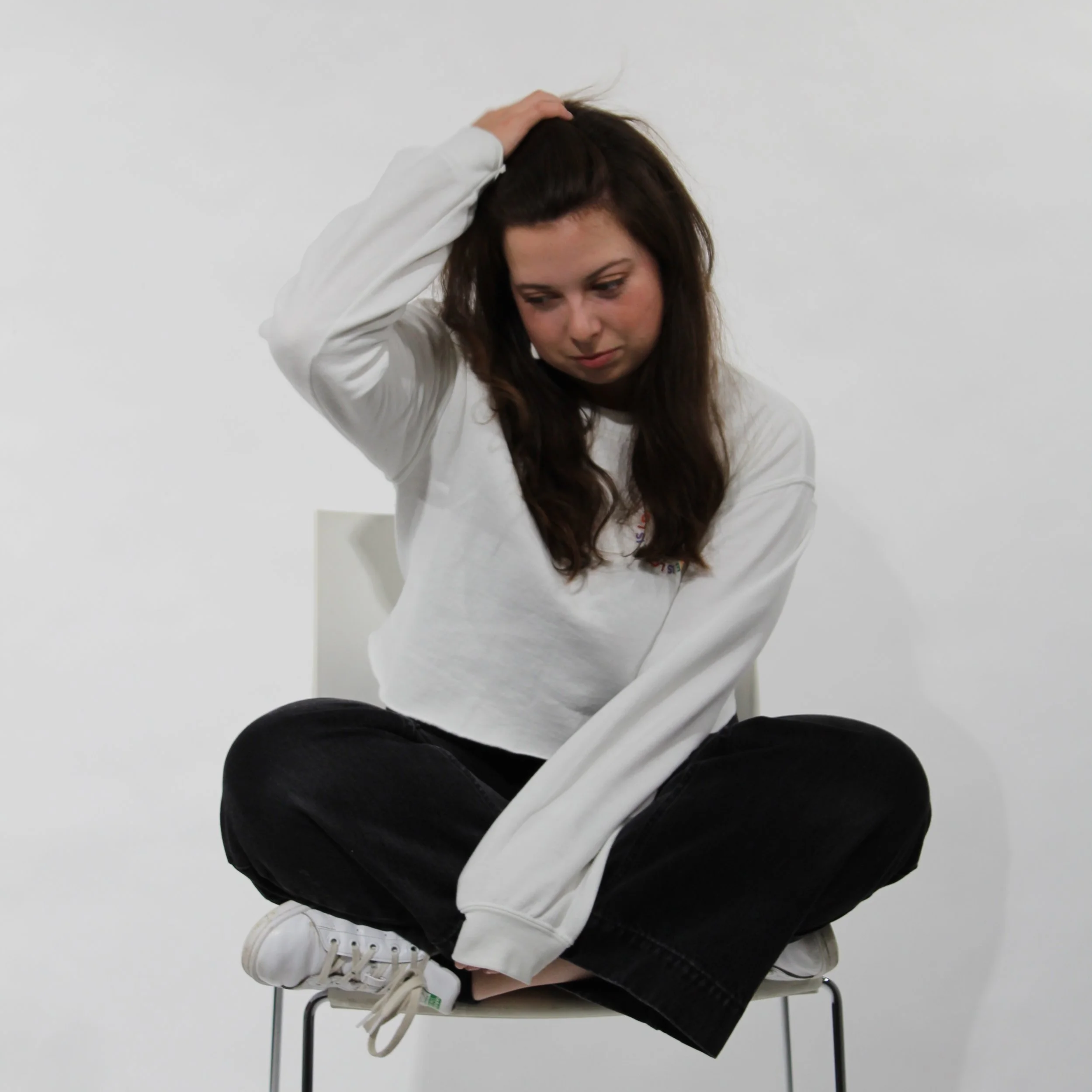 A young woman sitting cross-legged on a chair against a plain white background, holding her head with one hand and looking down.