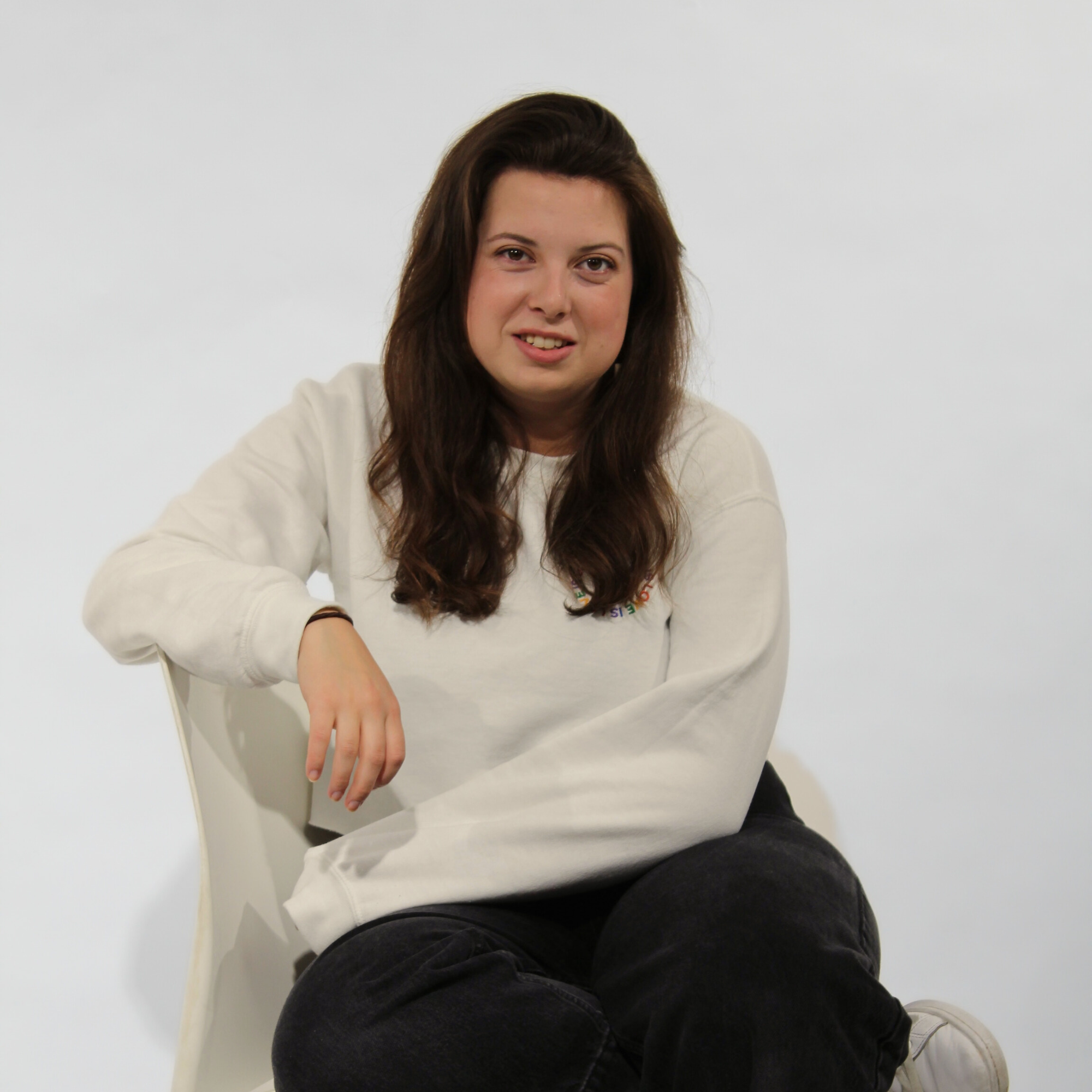 A young woman with long dark hair sitting on a white chair against a plain white background, wearing a cream sweatshirt and black pants.
