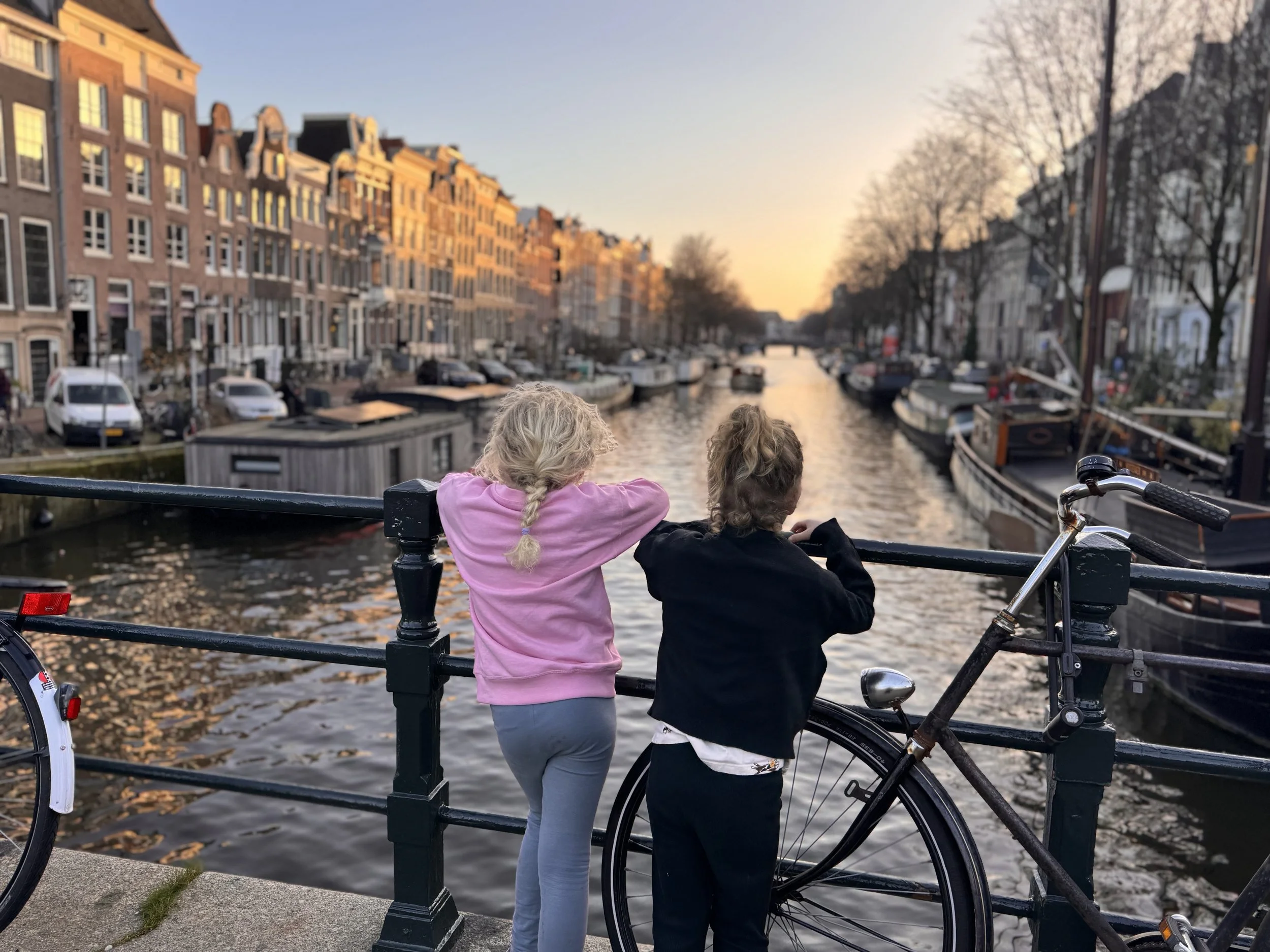 Two children, one with blonde hair and the other with brown hair, are leaning against a black metal railing on a bridge overlooking a canal with boats. The canal is lined with multi-story European-style buildings and trees without leaves. The sky is clear with the sun setting in the distance, casting a warm glow.