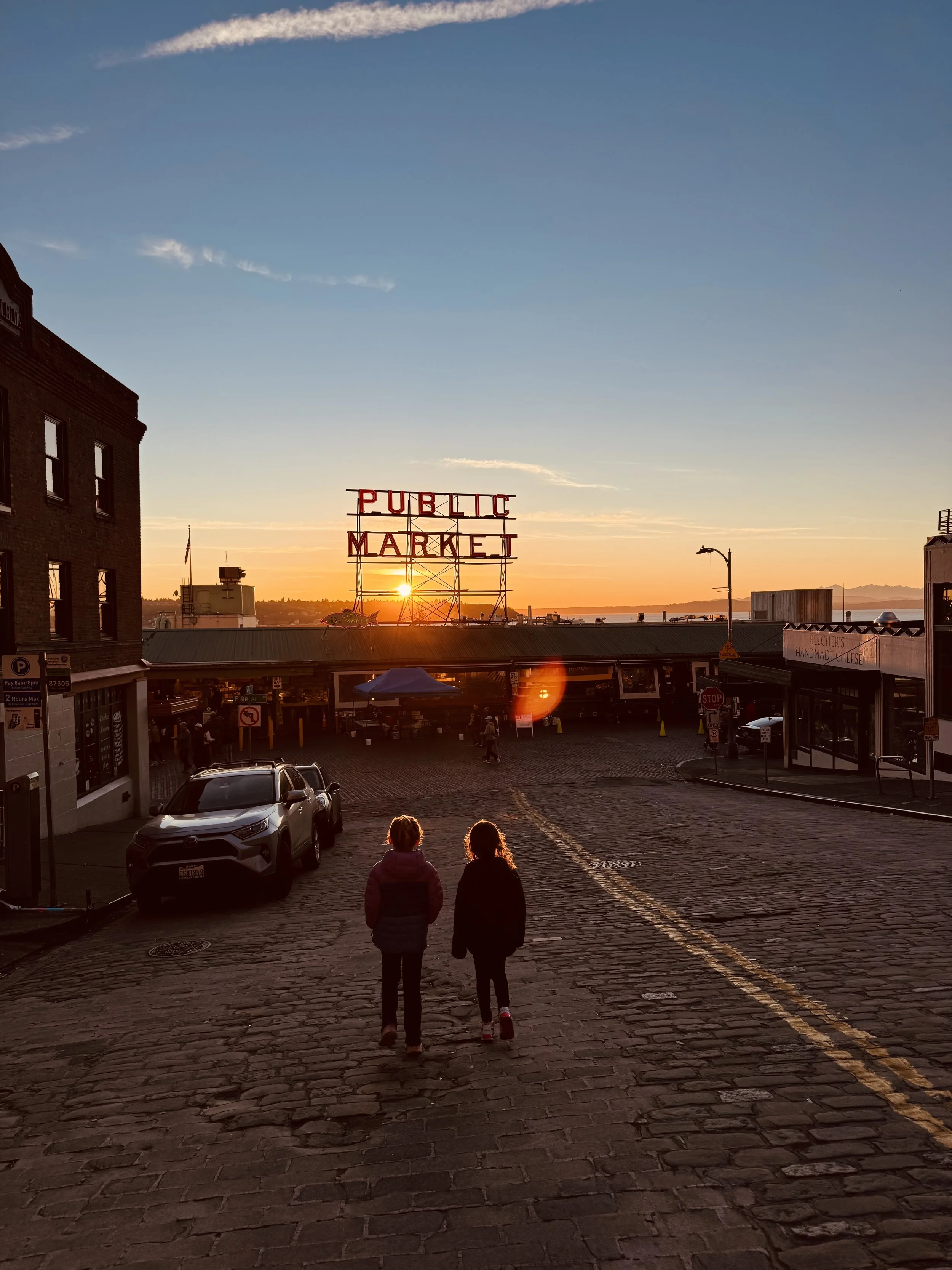 Two children walking on a Seattle street at sunset with Pike's 'PUBLIC MARKET' sign in the background.