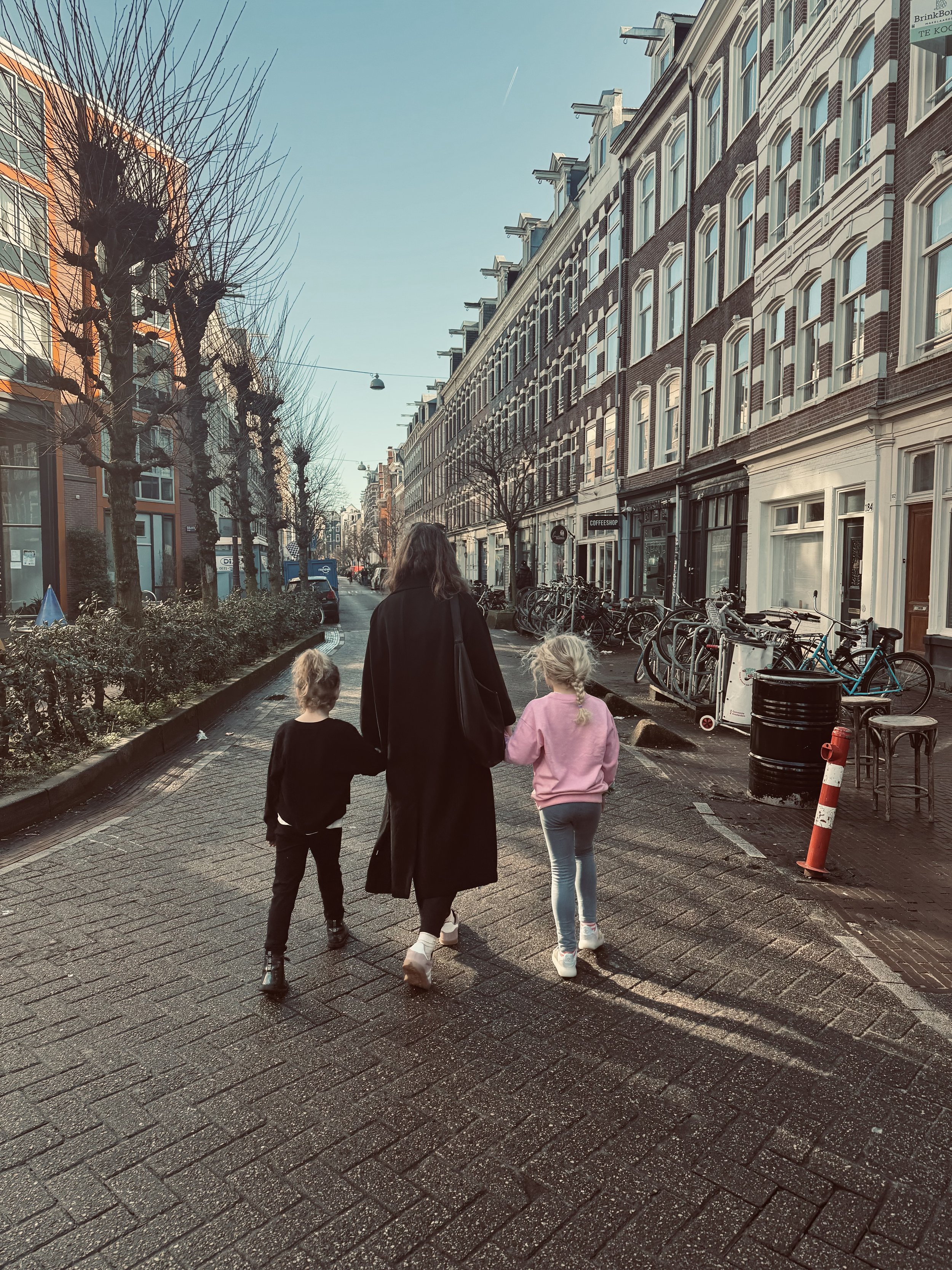 A woman walking with two young girls on a narrow Amsterdam  street. The street is lined with leafless trees, bicycles, and historic building facades.