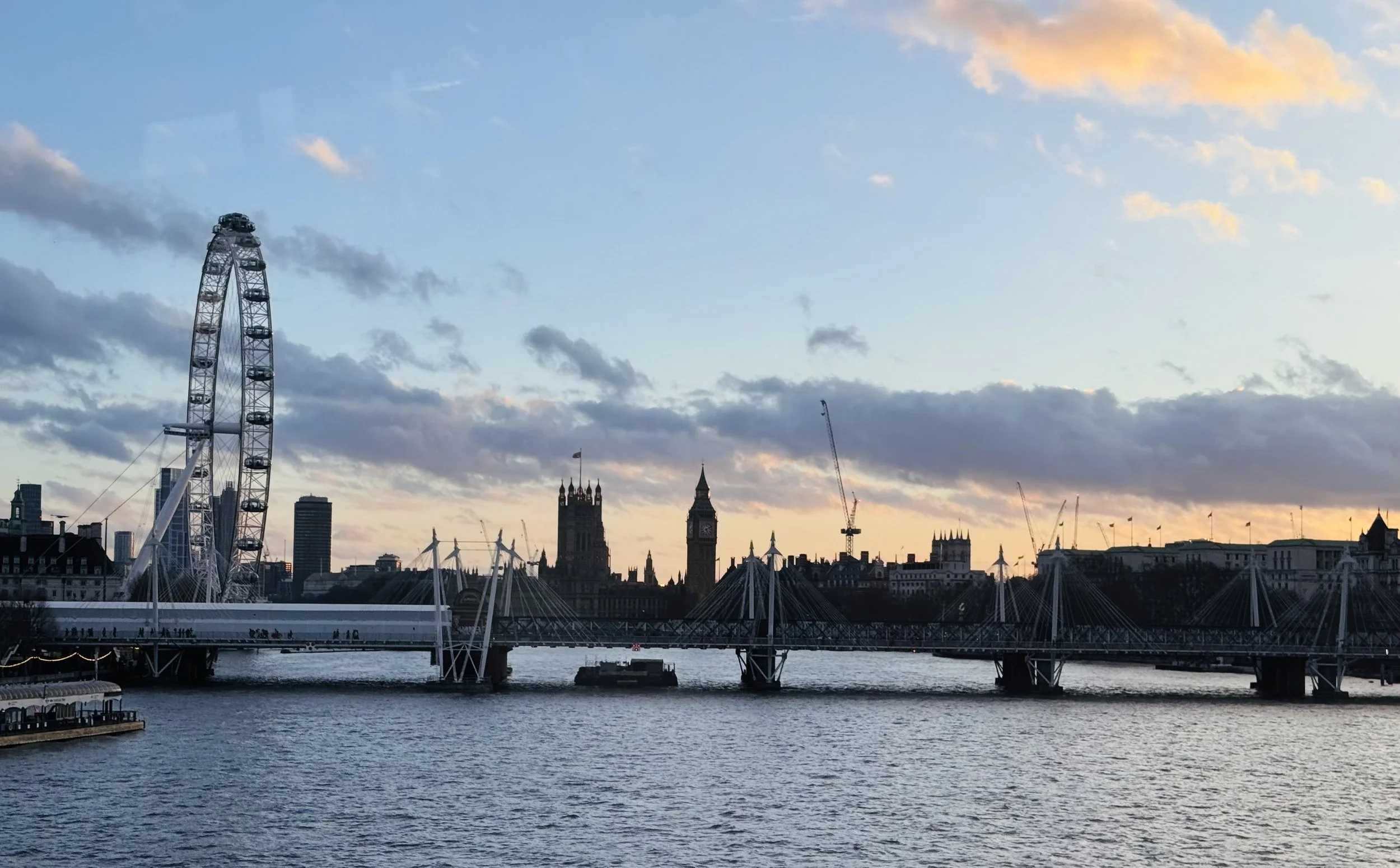 View of London skyline at sunset, featuring the London Eye, Big Ben, and the Houses of Parliament with cranes in the background.