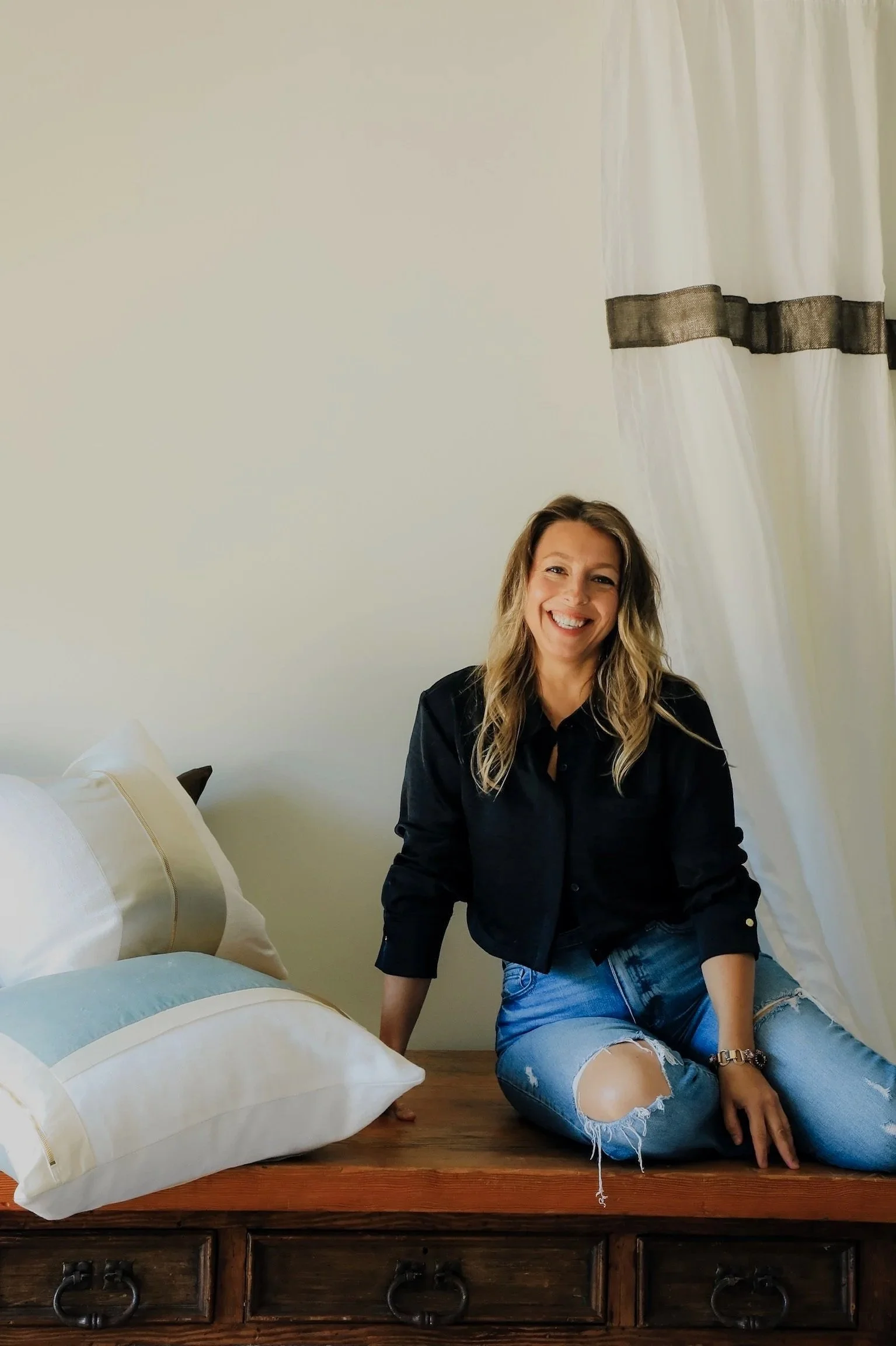 A woman with long blonde hair, wearing a black jacket and ripped jeans, sitting on a wooden bed frame, smiling, with pillows on the bed behind her and a white curtain with a black band on a window in the background. A passionate interior artist