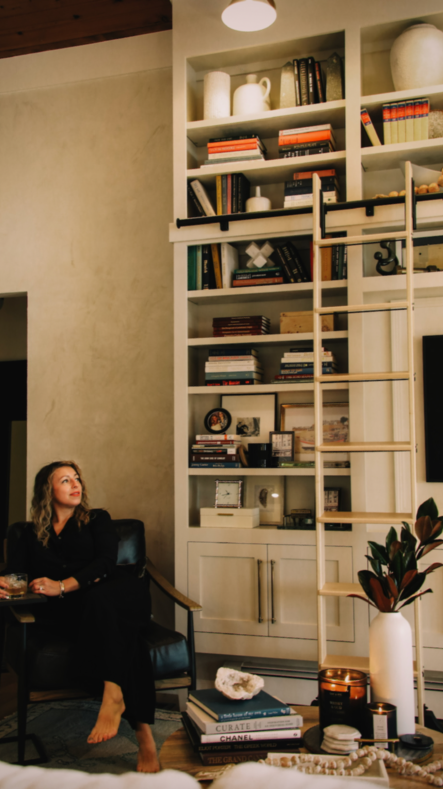 A woman sitting on a black chair in a living room, with a tall white bookshelf filled with books, decorative items, and framed photos. A wooden ladder leans against the bookshelf. There is a white vase with plants, candles, and a stack of books on a coffee table in front of her. The wall behind is beige with a wooden ceiling.