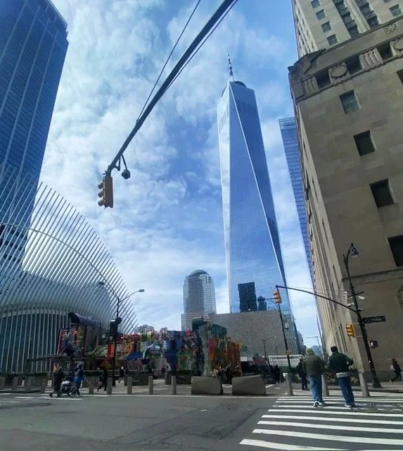 Downtown New York City street scene featuring One World Trade Center skyscraper with glass facade, nearby modern buildings, and people walking on the crosswalk.