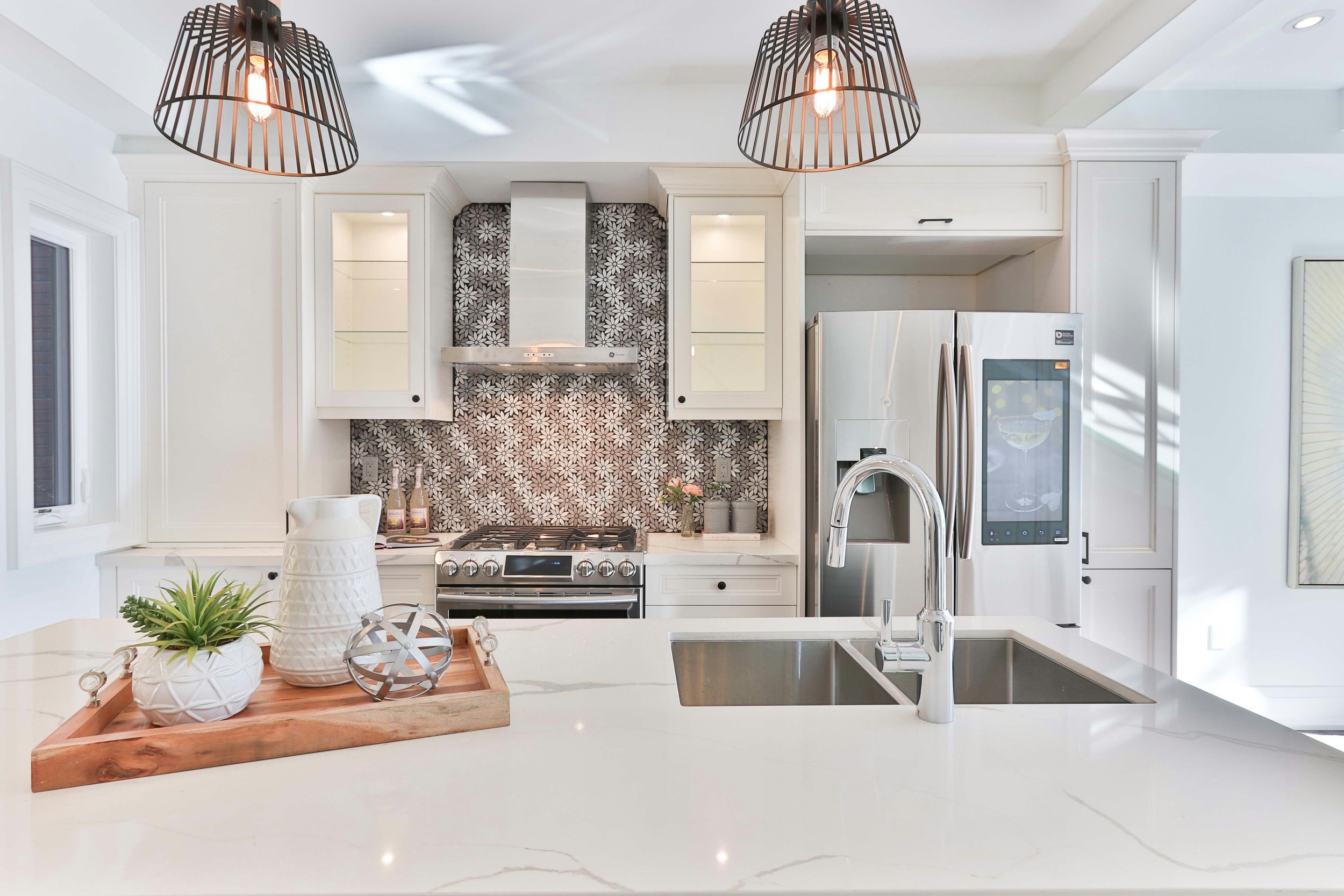 A modern kitchen with white cabinets, a stainless steel stove and refrigerator, black and white floral backsplash, decorative lighting fixtures, and a kitchen island with potted plants and decor.
