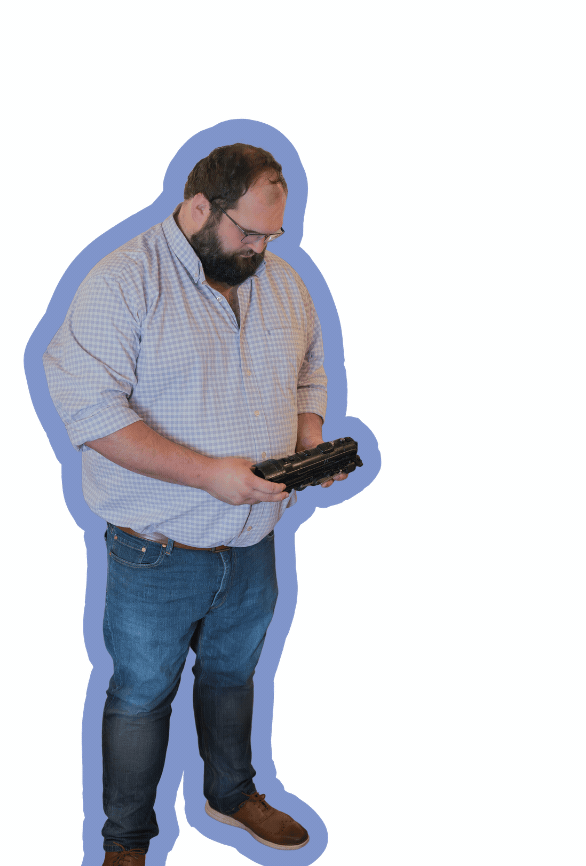 A man with glasses, a beard, wearing a checkered shirt and jeans, is looking down at a toy train, standing on a white background with a blue shadow outline.
