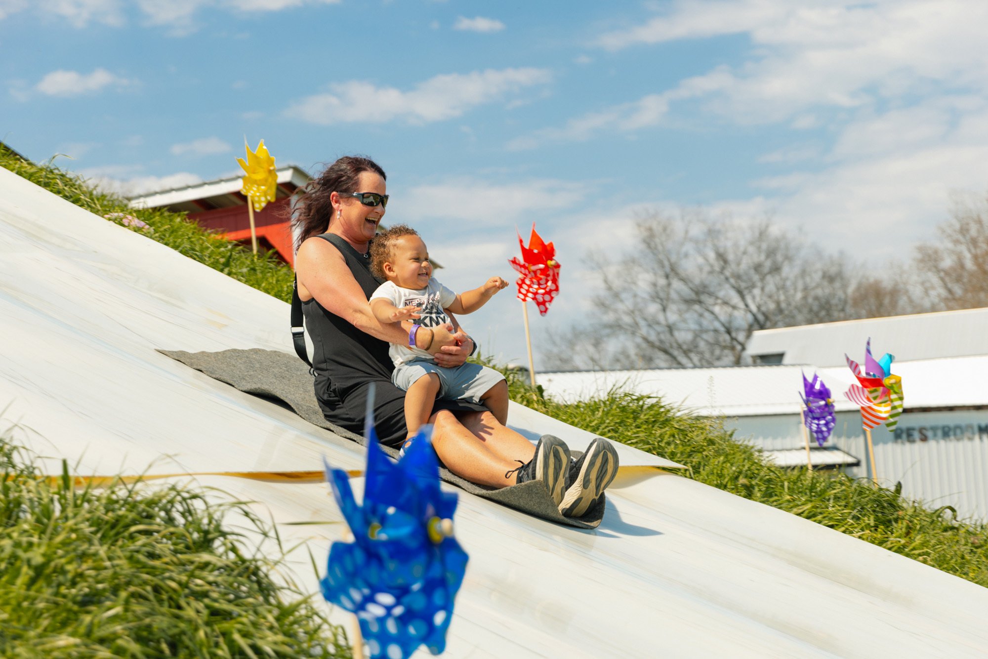 A woman with sunglasses and a child sitting on a slide outdoors during daytime at Donnell Century Farms, with colorful wind spinners around them and a building in the background.