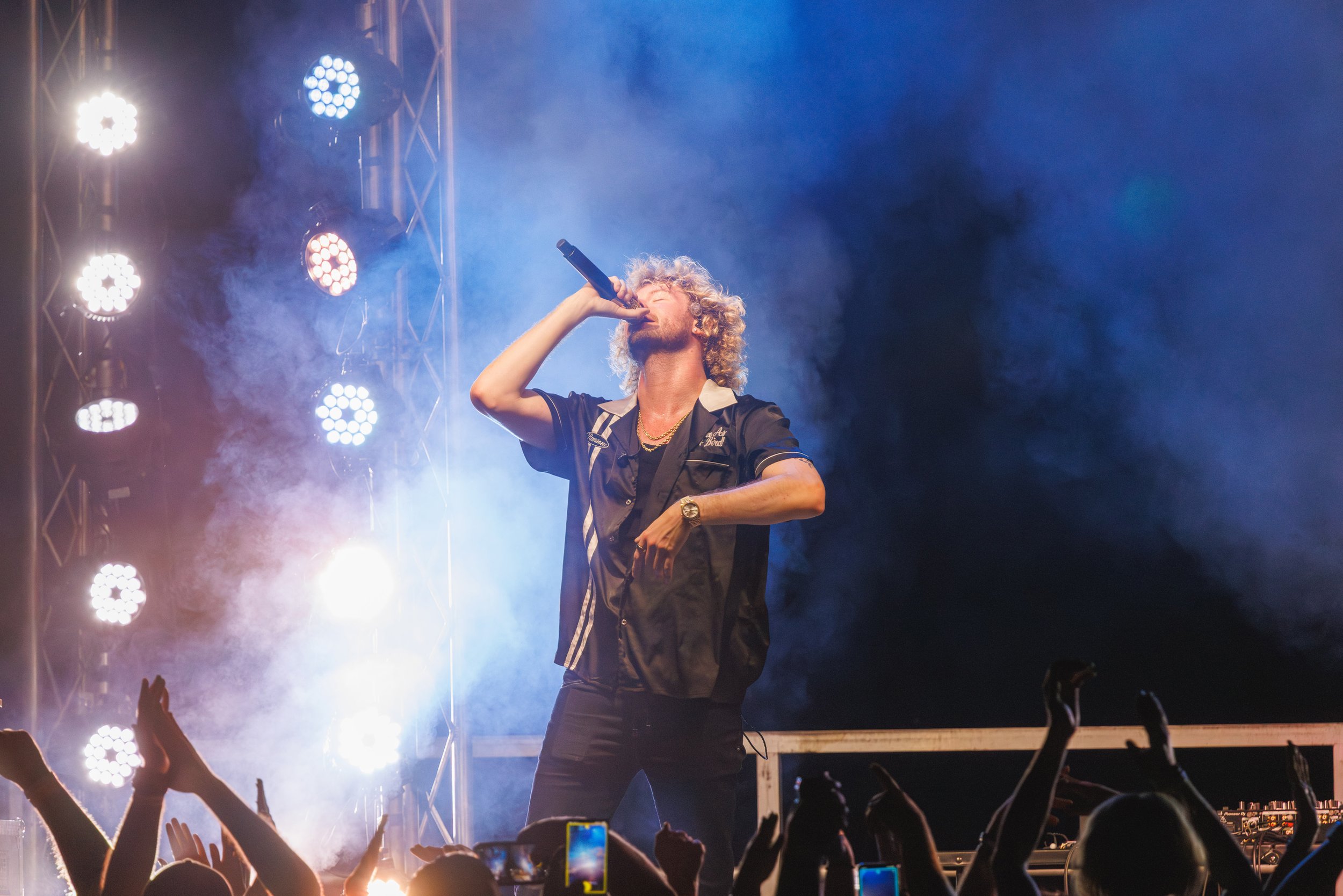 Male rapper, Yung Gravy, singing into a microphone on stage during a concert with bright stage lights and an enthusiastic crowd in front.