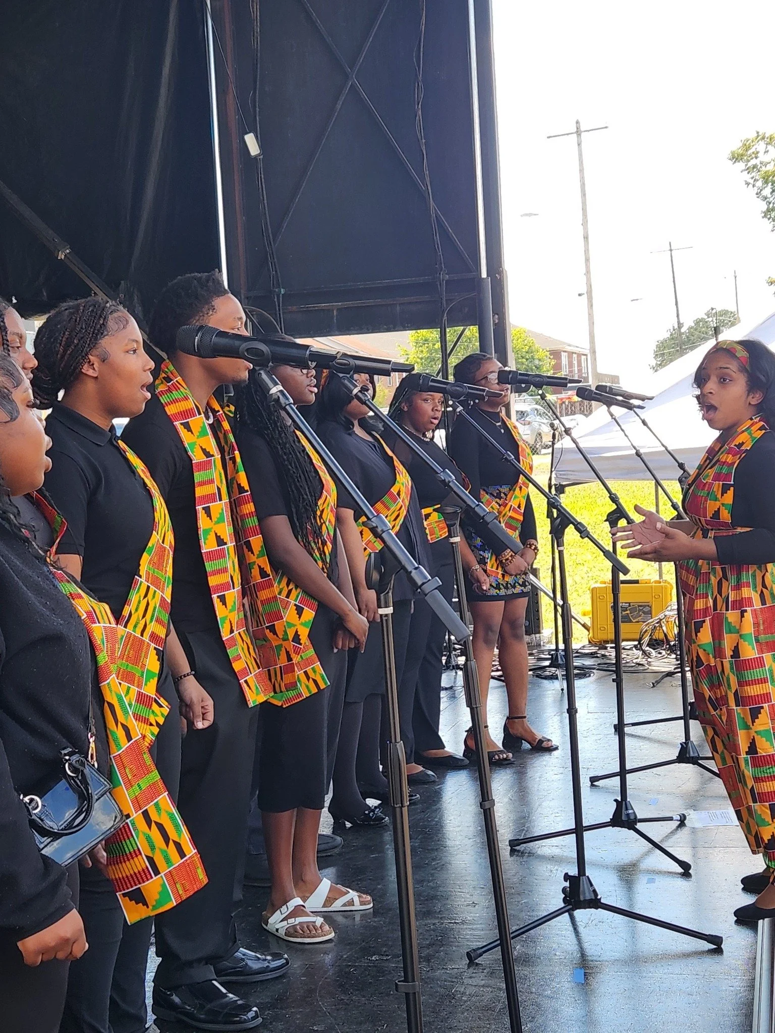 A group of women singing together on an outdoor stage, wearing matching black shirts and colorful kente scarves, with microphones in front of them.