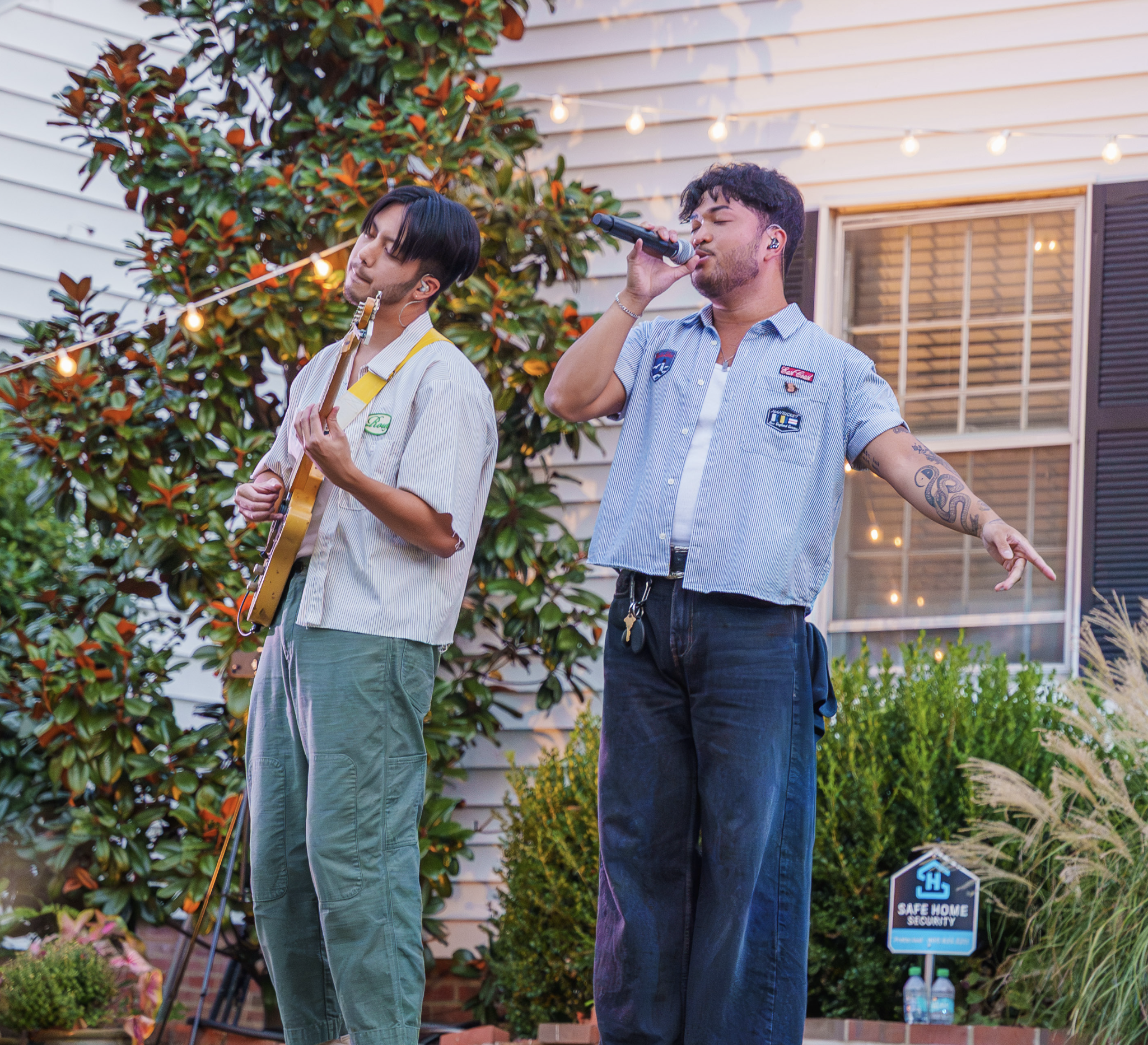 Two young men performing outdoors at sunset, one singing into a microphone and the other playing guitar, with string lights overhead and garden plants in the background.