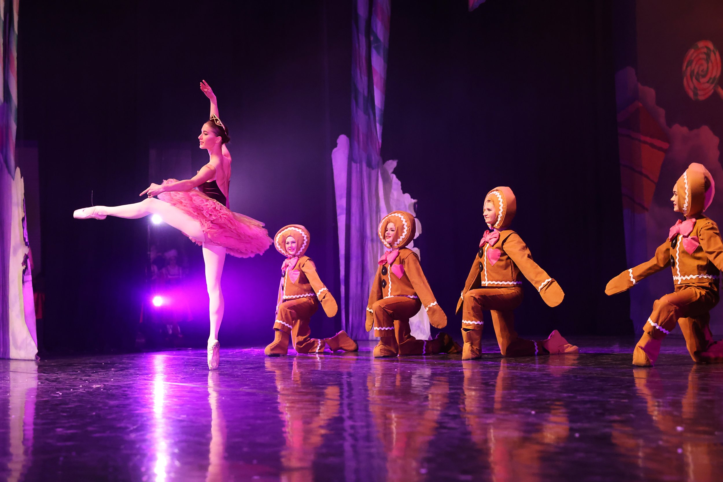 A ballet dancer performs on stage in The Nutcracker wearing a pink tutu, framed by a decorated theater setting. Four dancers dressed as gingerbread cookies kneel in a line behind her.