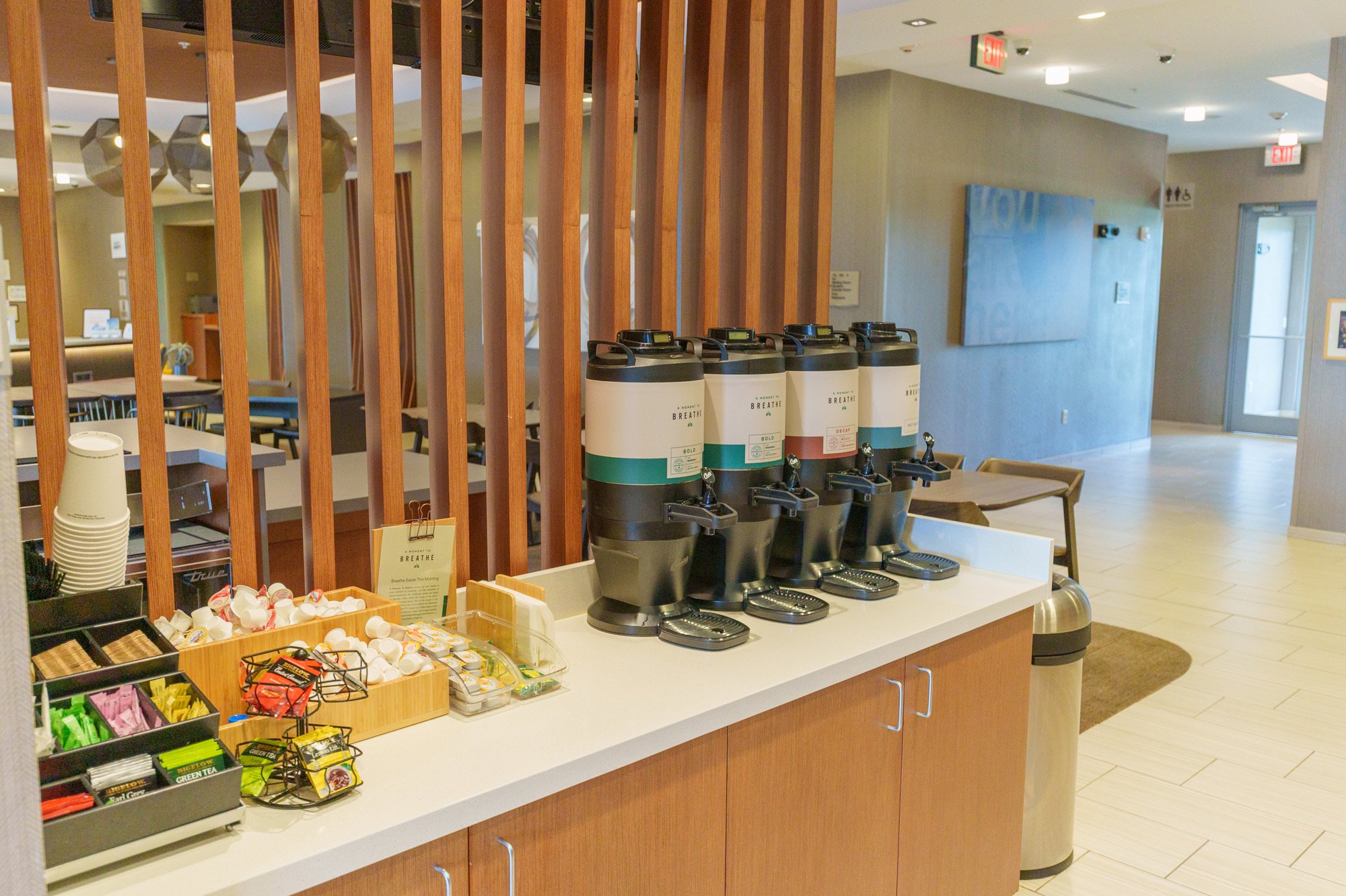 Hotel coffee station with multiple coffee dispensers, a selection of tea bags, and condiments in a cafe or lounge area.