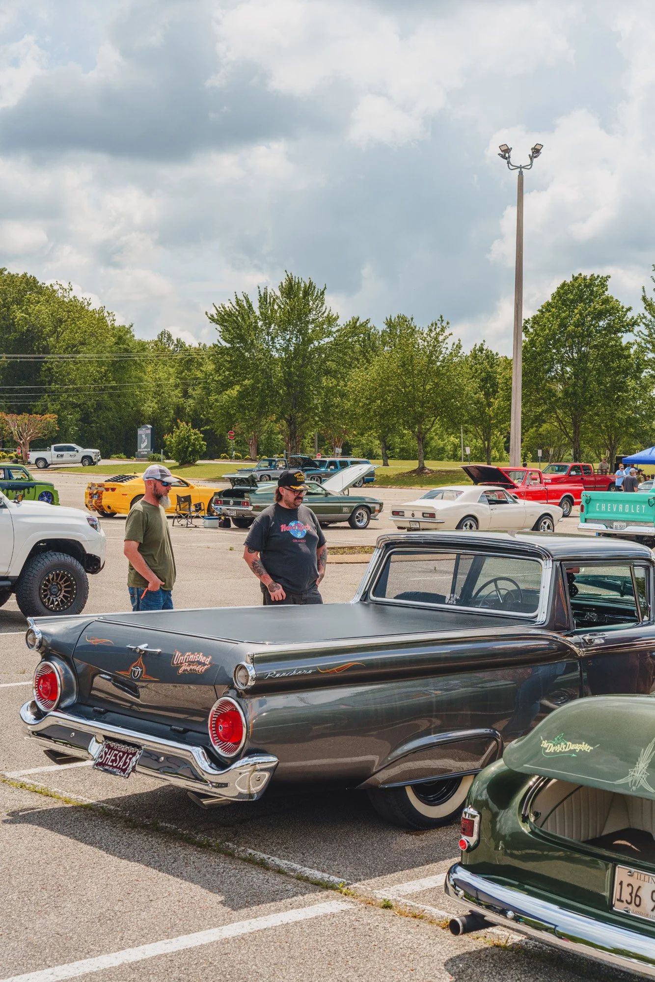 Classic cars parked in a lot at a car show with people walking around, trees, and a cloudy sky in the background.