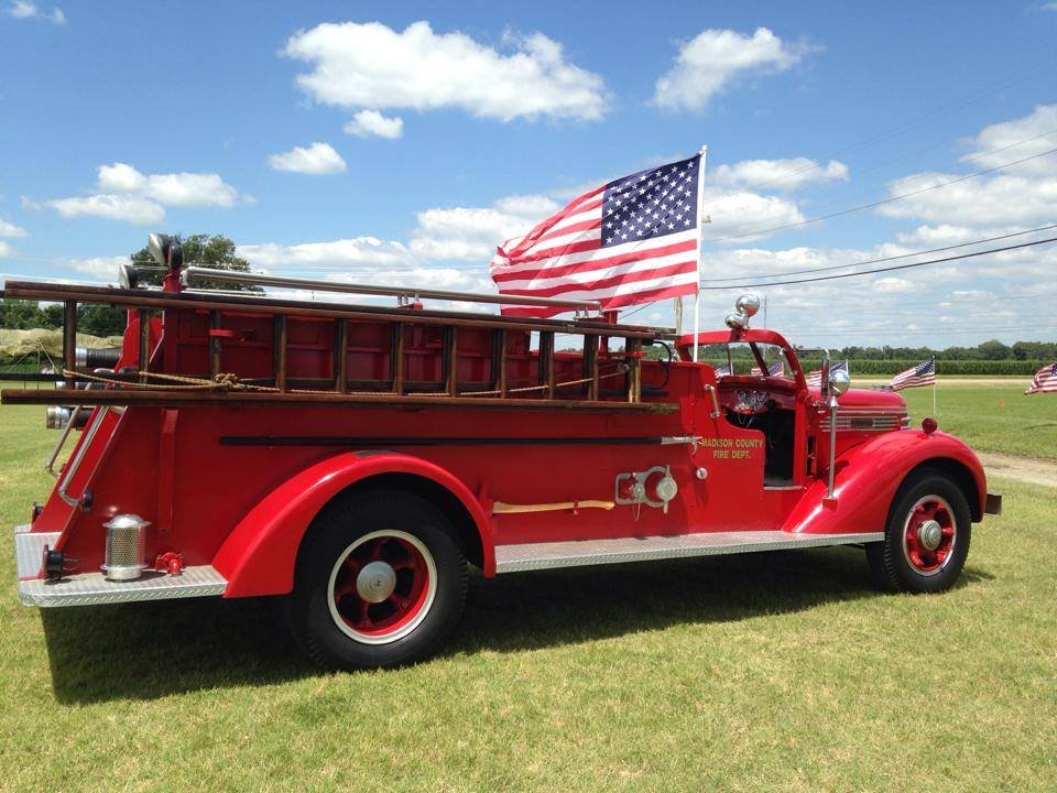 Red vintage fire truck with an American flag attached, parked on a grassy field under a partly cloudy sky.