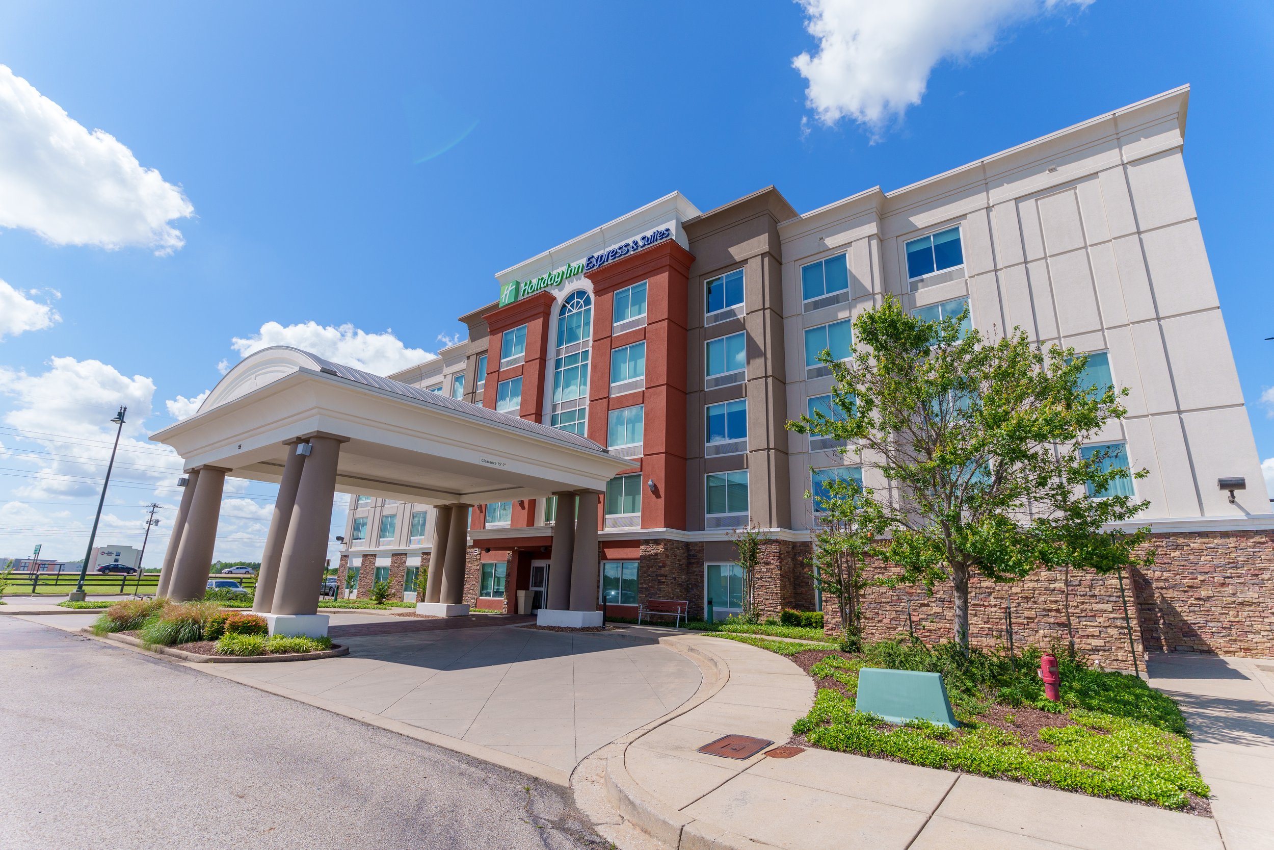 Exterior of a Holiday Inn Express & Suites hotel under a bright blue sky with some clouds, featuring a decorated entrance with a large archway and a parking lot with several cars.