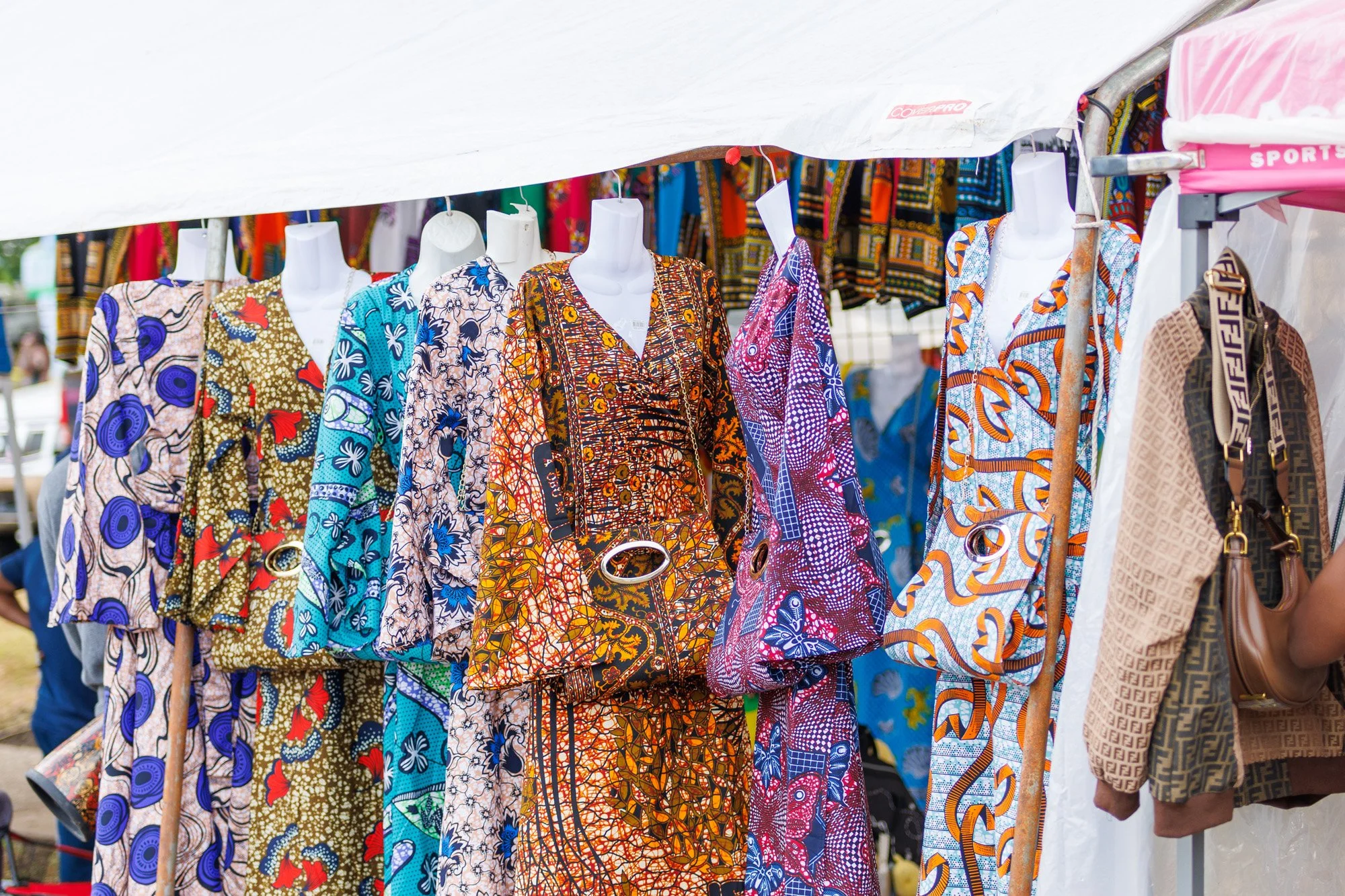 Vendors display colorful patterned clothing and accessories at an outdoor market stall with a white canopy.
