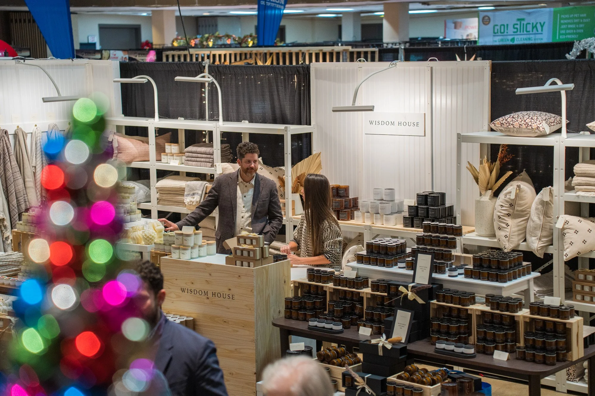 Indoor market booth labeled 'WISDOM HOUSE' selling home decor, textiles, and candles, with a man and woman browsing and a colorful blurred Christmas tree in the foreground.