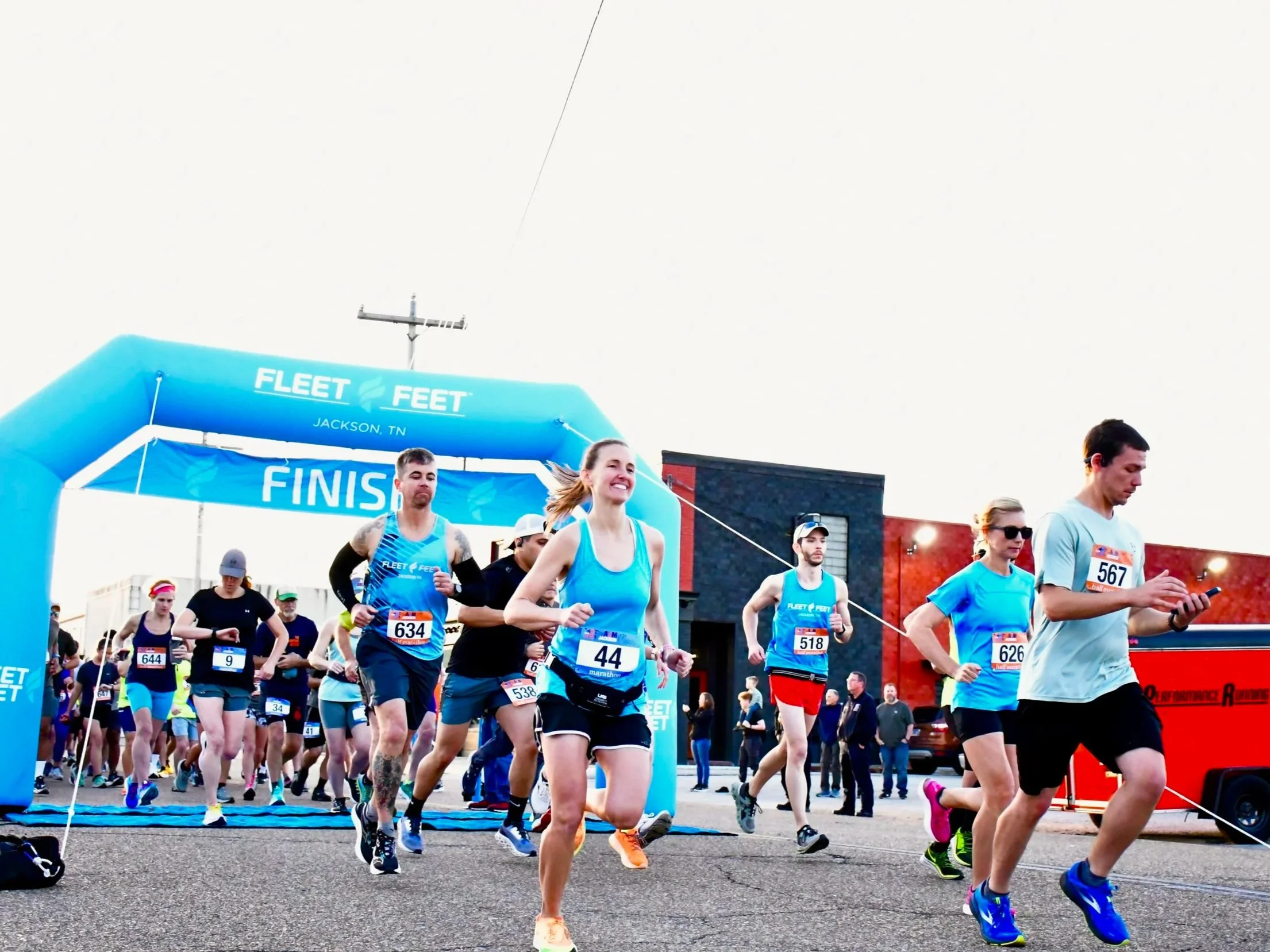 Group of marathon runners crossing the finish line during a race in Jackson, Tennessee, with many wearing matching blue shirts.